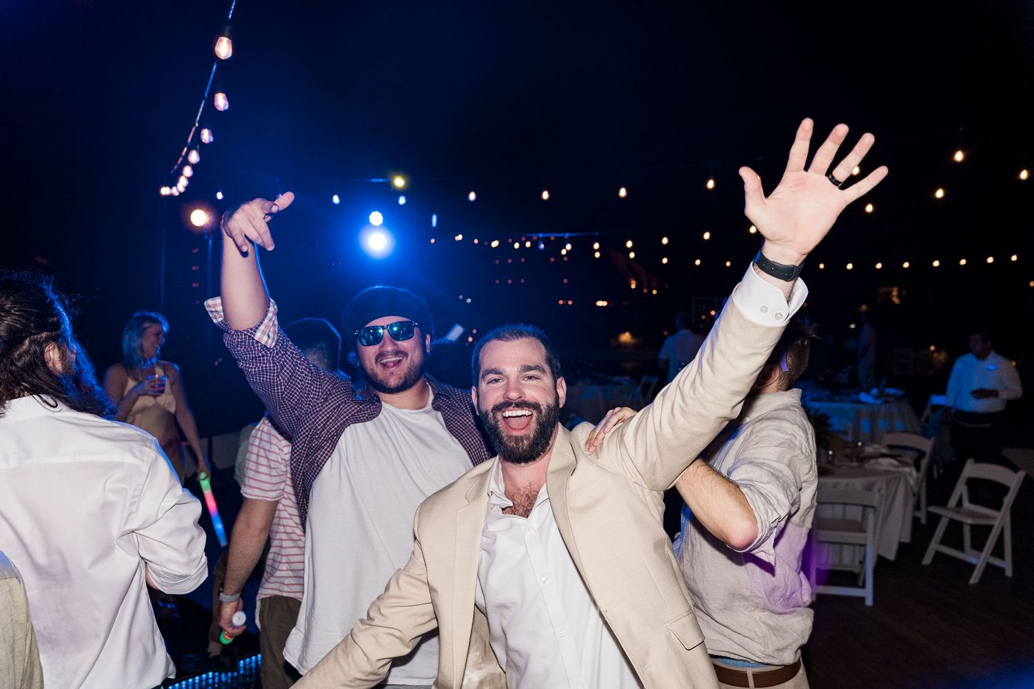 Two men dancing joyfully at an outdoor evening wedding reception under string lights.