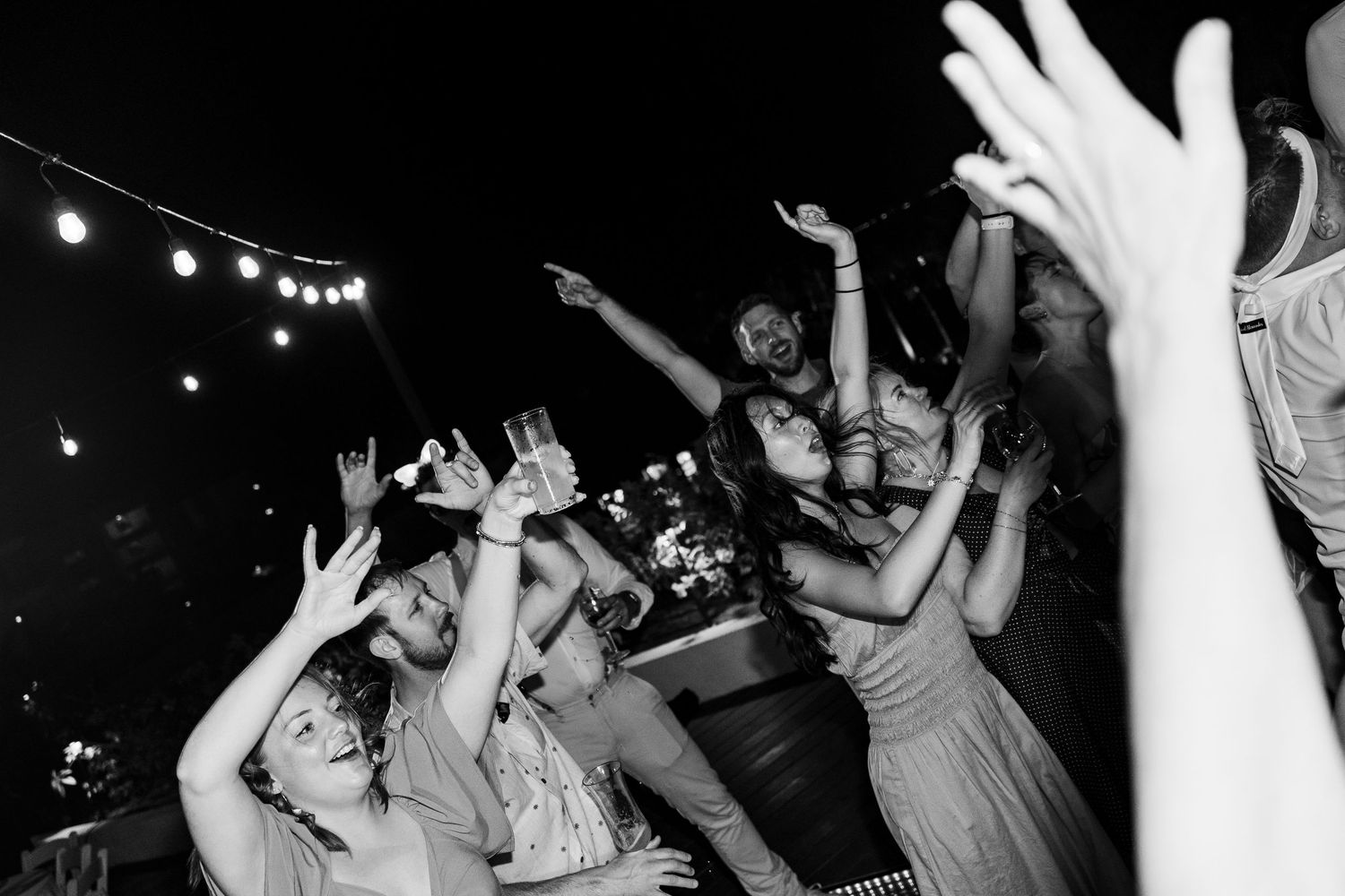 Black and white photo of excited crowd dancing with hands raised at an outdoor evening party or wedding reception.