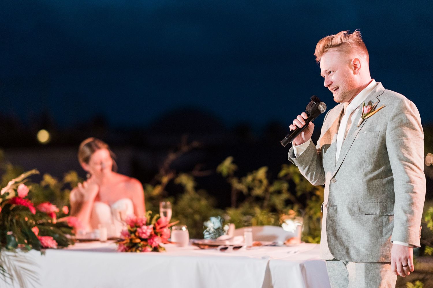 Groom in light gray suit gives wedding speech with microphone while bride smiles at outdoor evening reception.