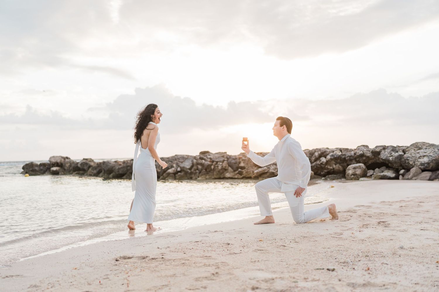 Man proposes on one knee to woman on a sandy beach at sunset, both dressed in white, with rocky shoreline backdrop.