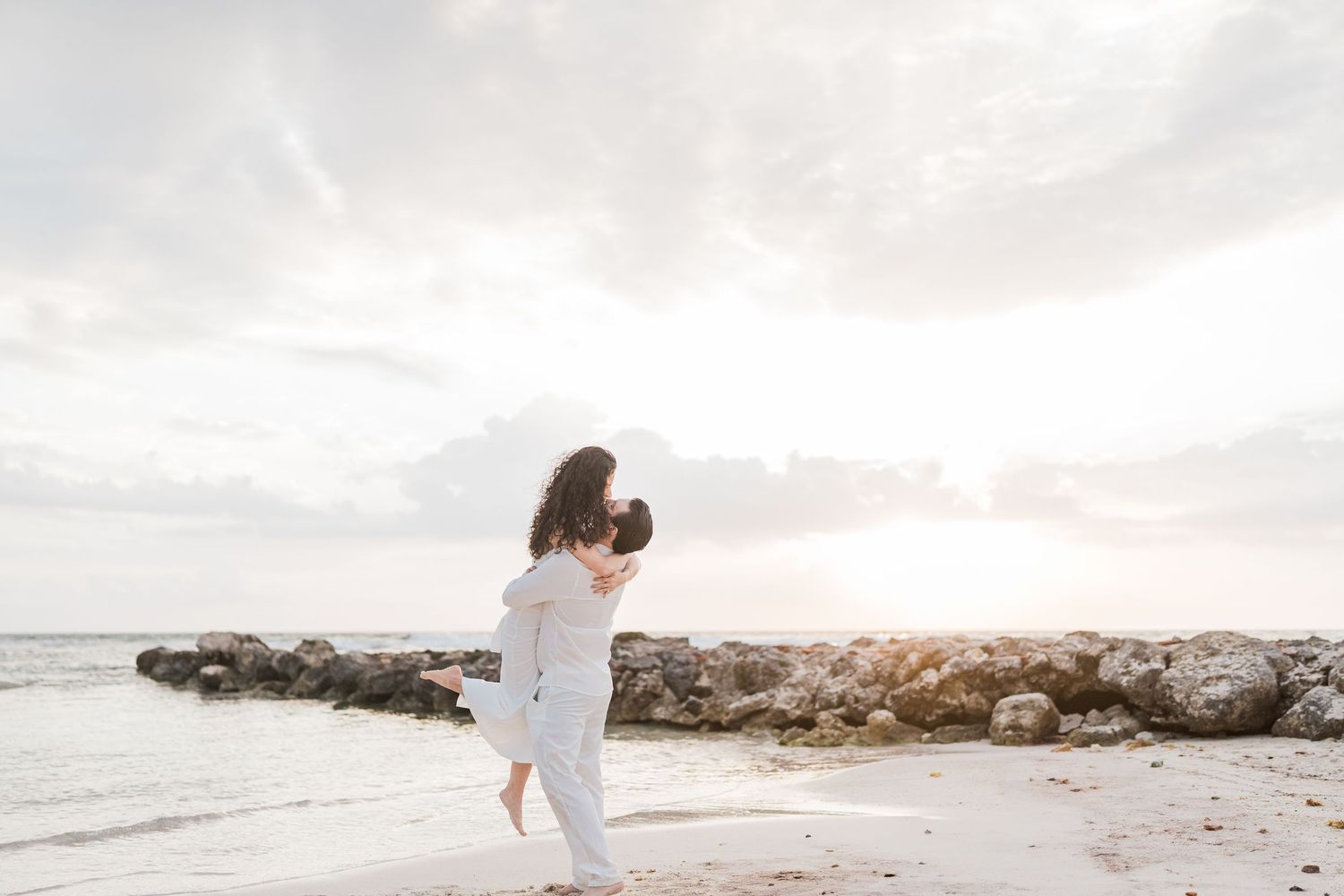 Couple embracing on a sandy beach at sunset with rocky shoreline and dramatic cloudy sky in background.