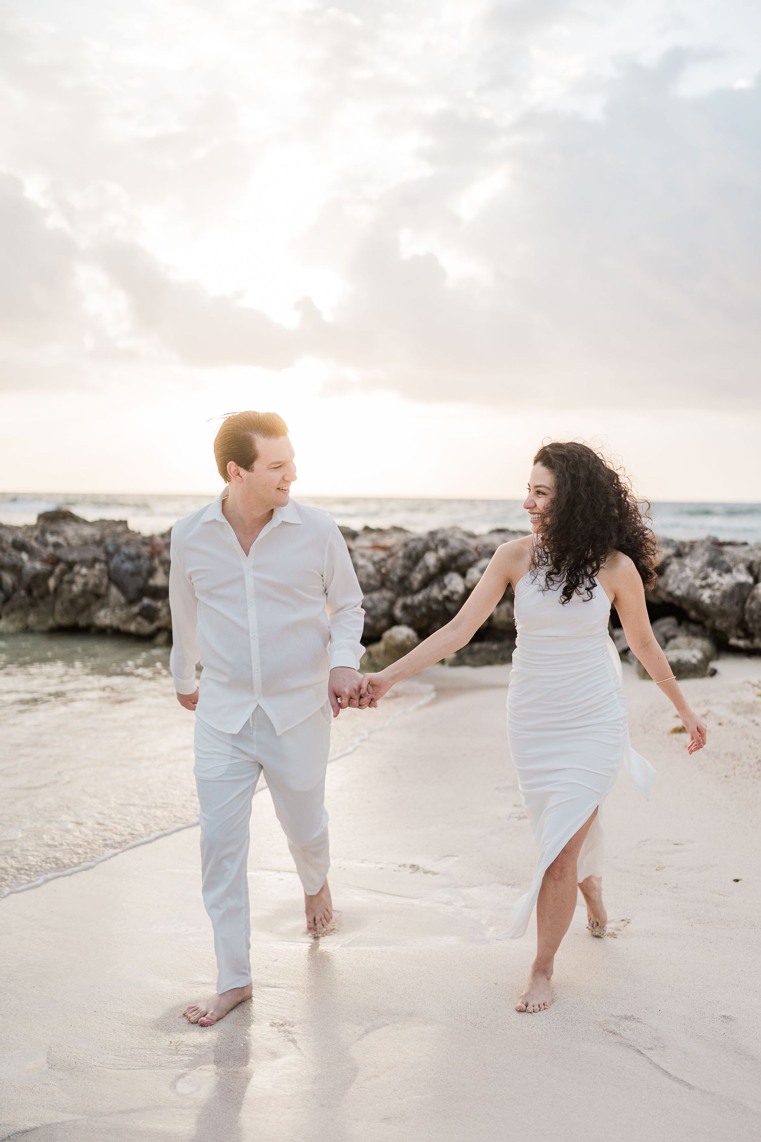A couple in white outfits walks hand-in-hand on a beach at sunset, smiling at each other near rocky shores.