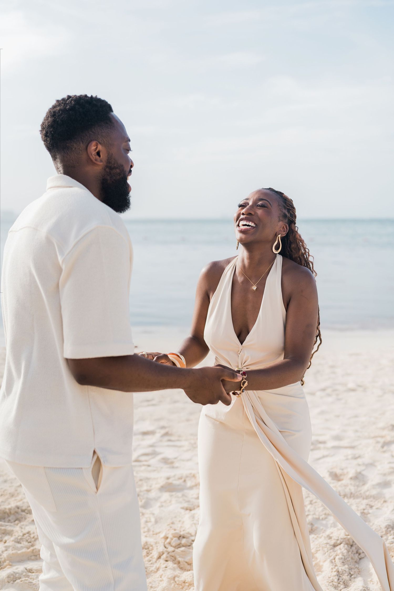 A joyful couple in white attire holds hands on a sunny beach, sharing a romantic moment by the ocean.