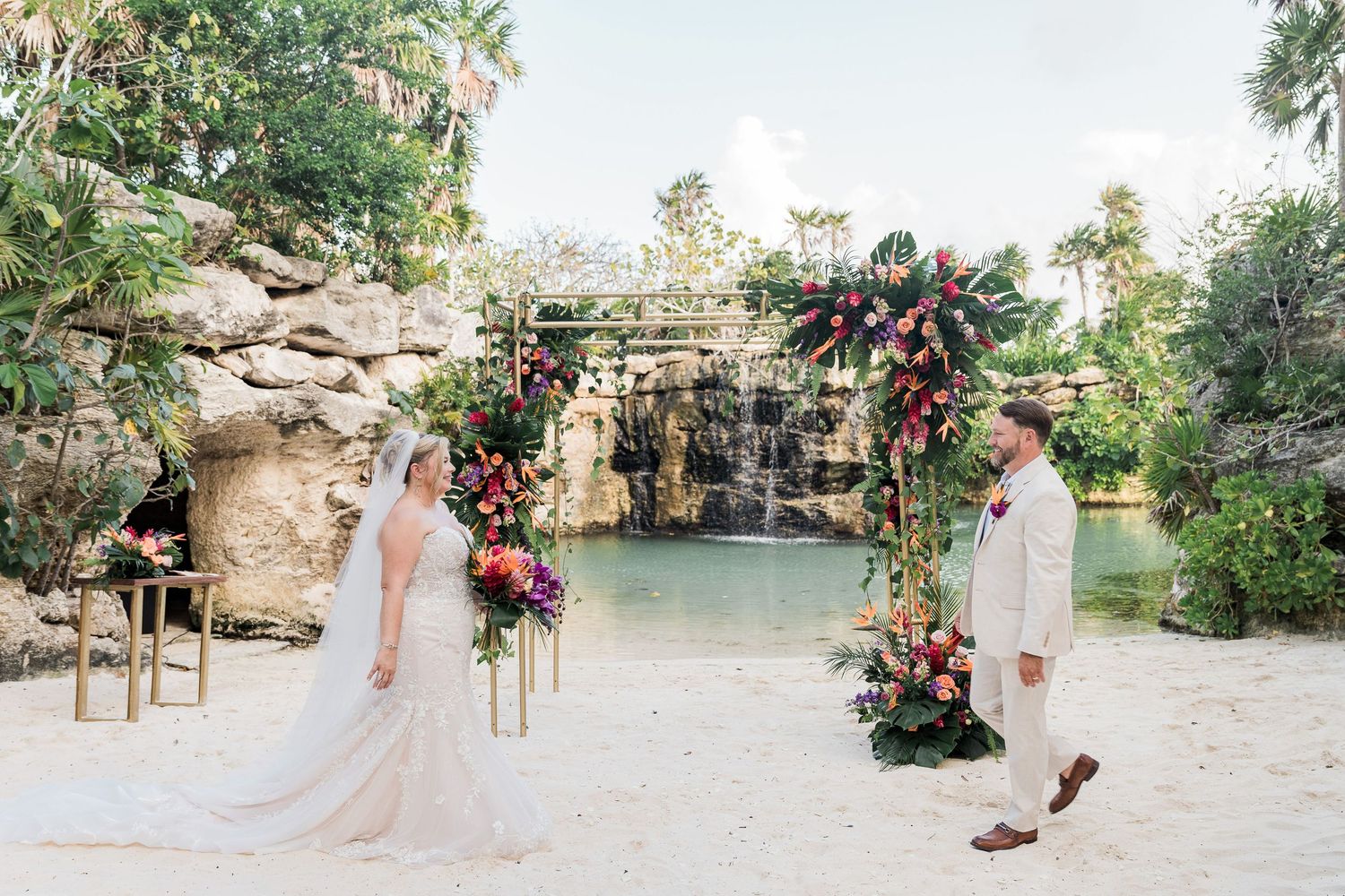 Bride and groom exchange vows under floral arch on sandy beach with tropical waterfall backdrop.