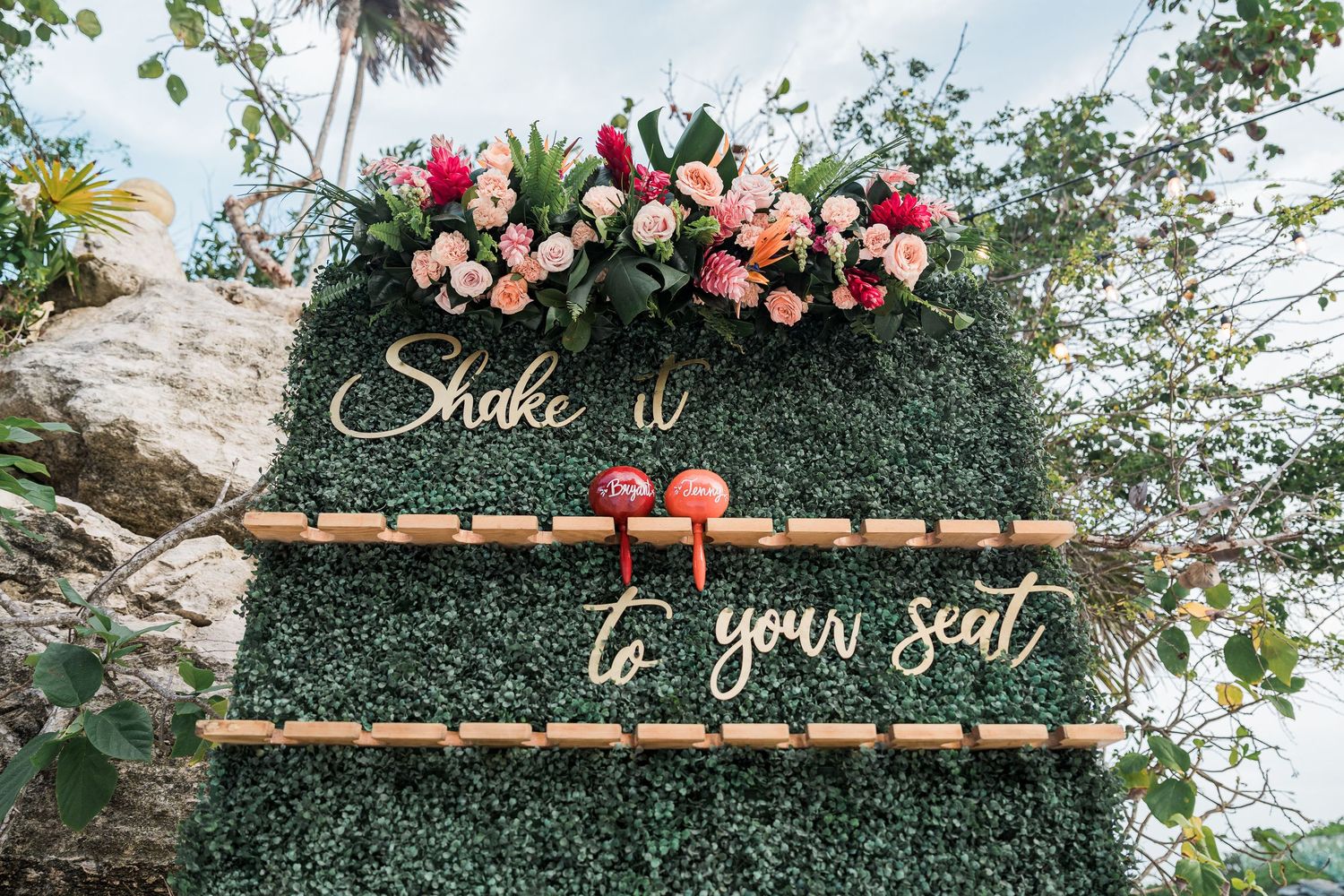 Green hedge wall wedding sign reading Shake it to your seat with pink floral arrangement and bamboo escort card holders.