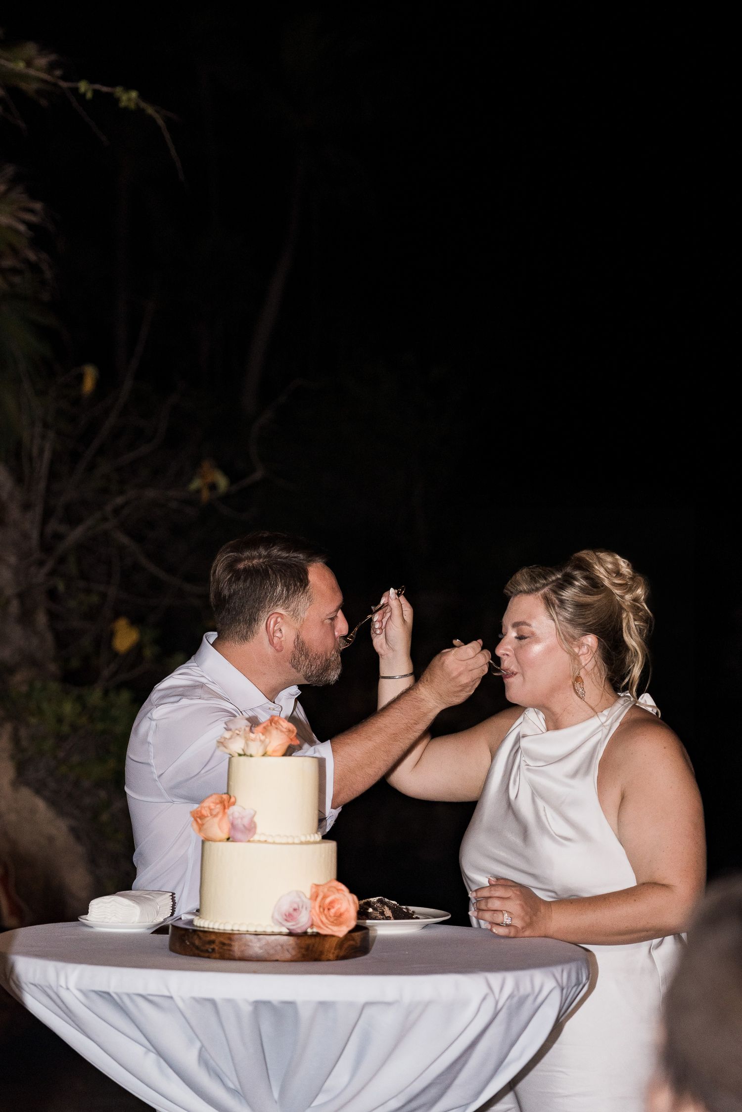 Bride and groom feeding each other wedding cake at outdoor nighttime reception with white tiered cake decorated with peach roses.