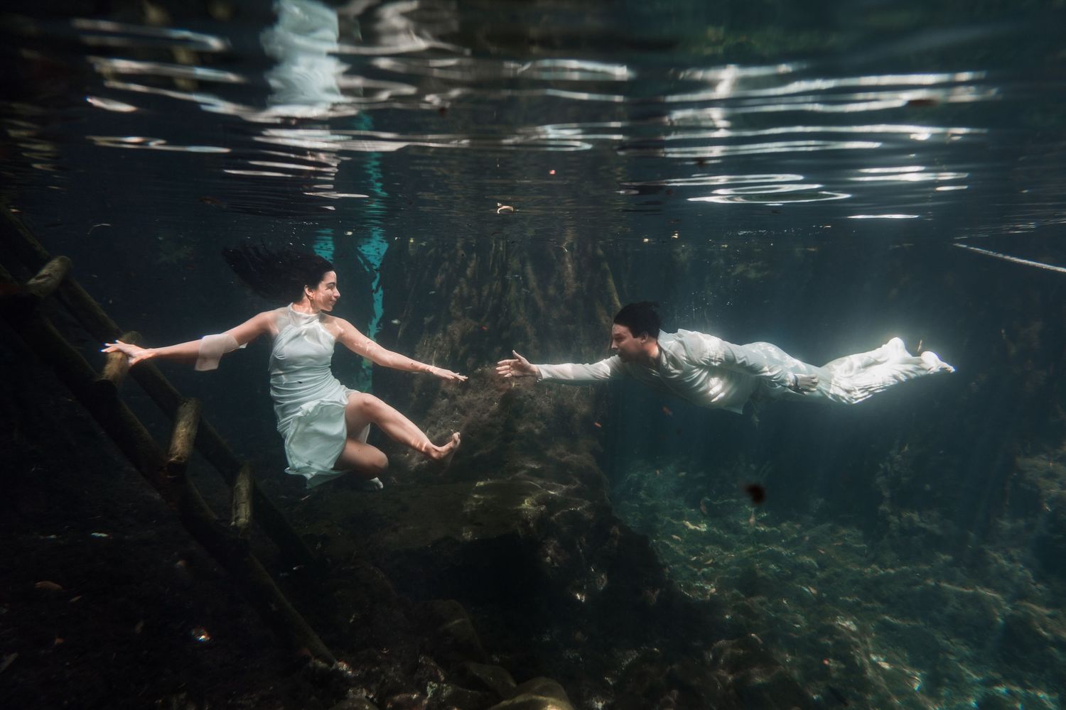 Couple in white attire holding hands while swimming underwater in a clear cenote during an underwater wedding photoshoot.