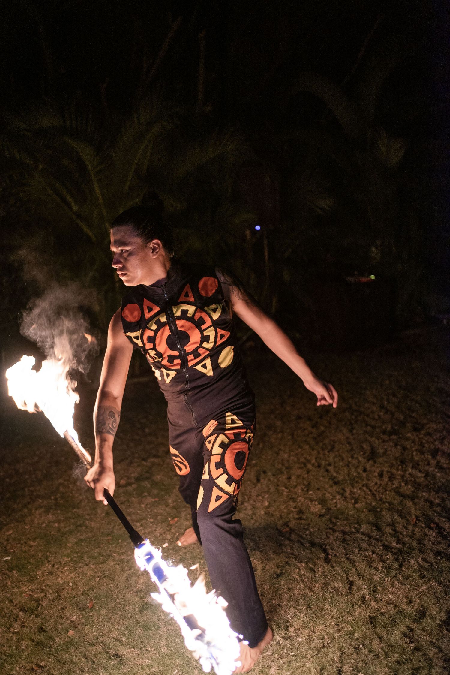 Fire dancer in tribal costume performing with flaming batons at an outdoor nighttime wedding celebration.