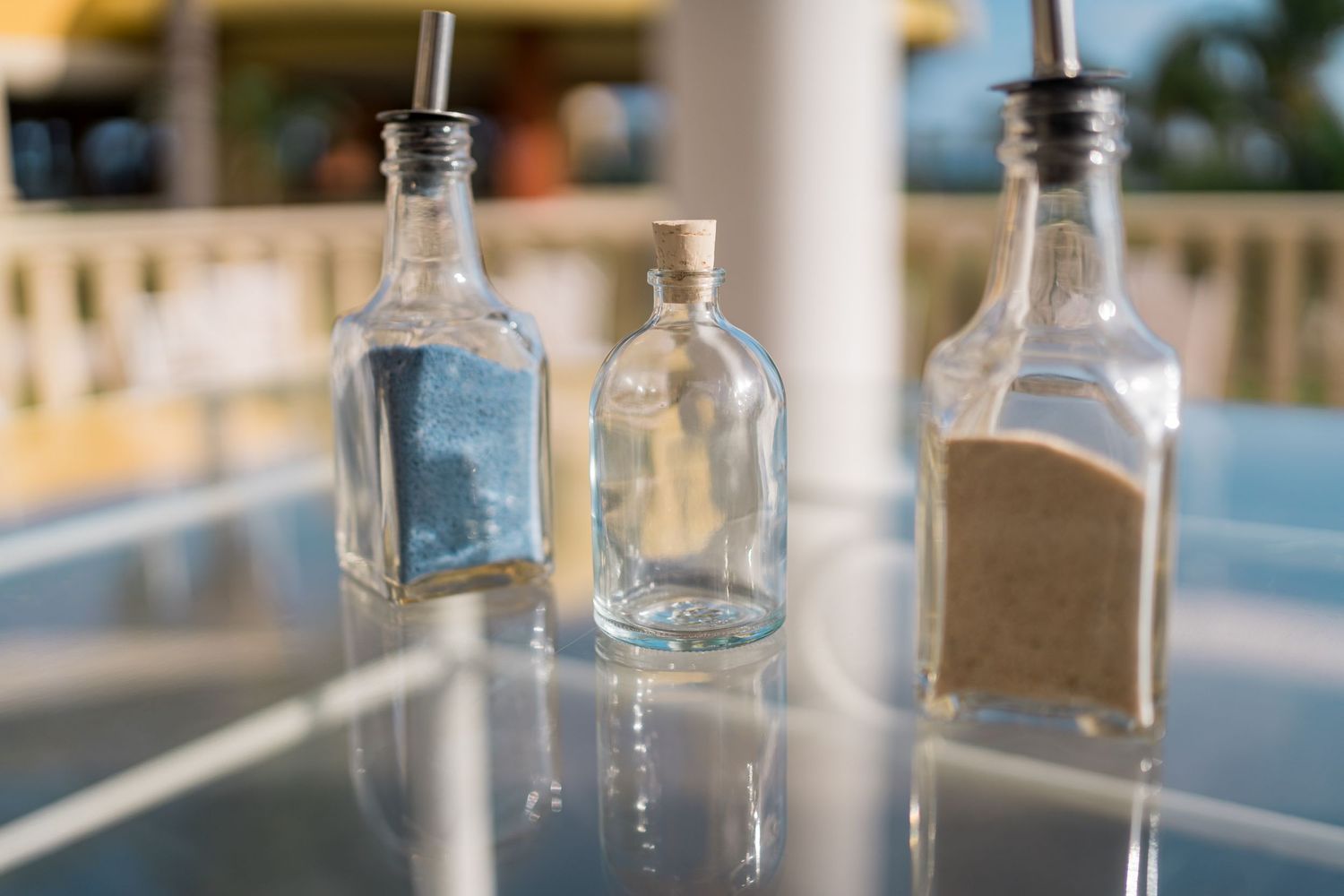 Three decorative glass bottles with sand and liquid on a glass table at an outdoor wedding venue.
