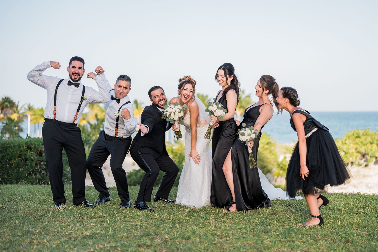 Energetic wedding party poses playfully on green lawn with ocean backdrop, bridesmaids in black gowns.
