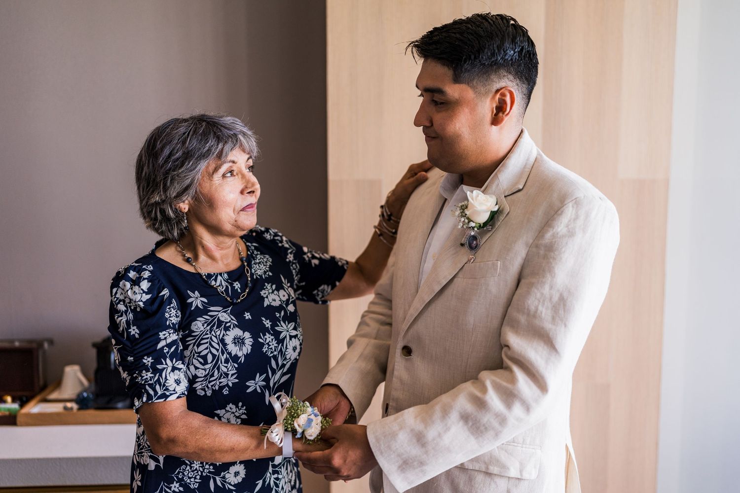 Groom in beige suit shares tender moment with mother during wedding preparations.