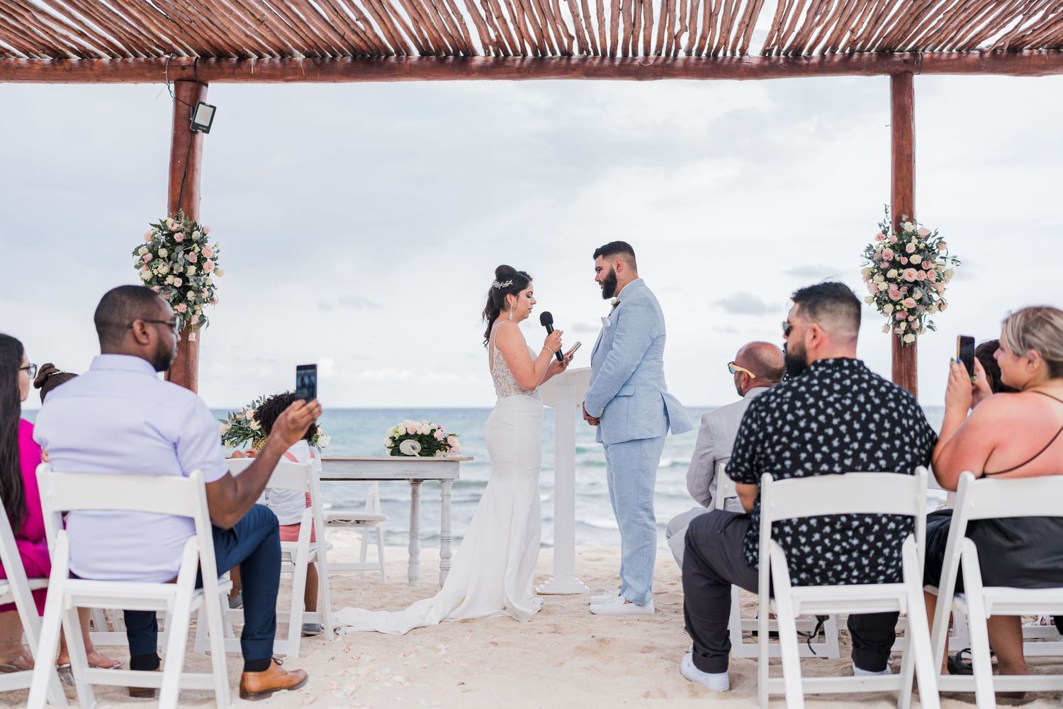 Wedding couple exchanges vows under thatched pergola on sandy beach while guests watch ceremony.