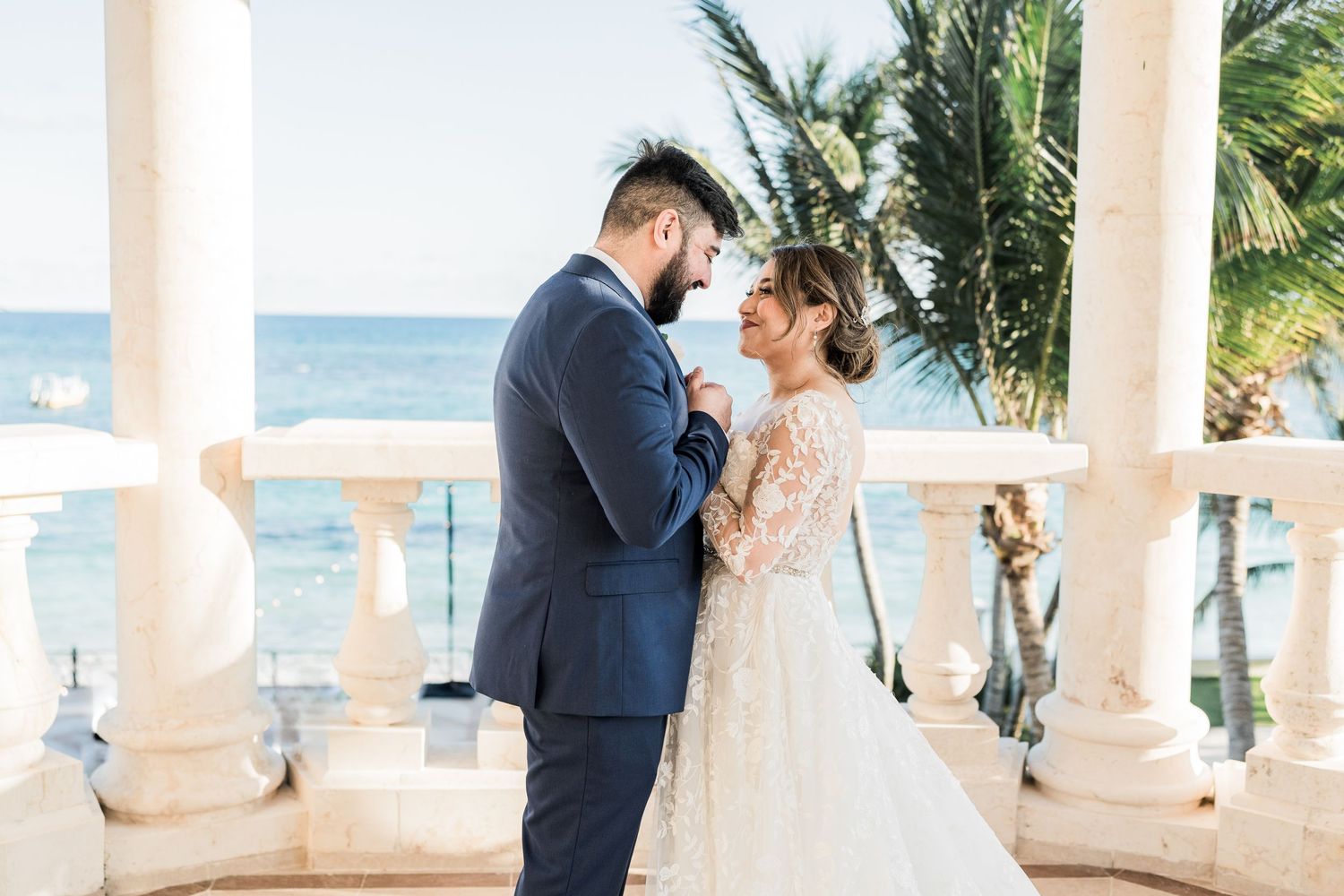 Bride in lace gown and groom in navy suit sharing tender moment on balcony overlooking tropical ocean.