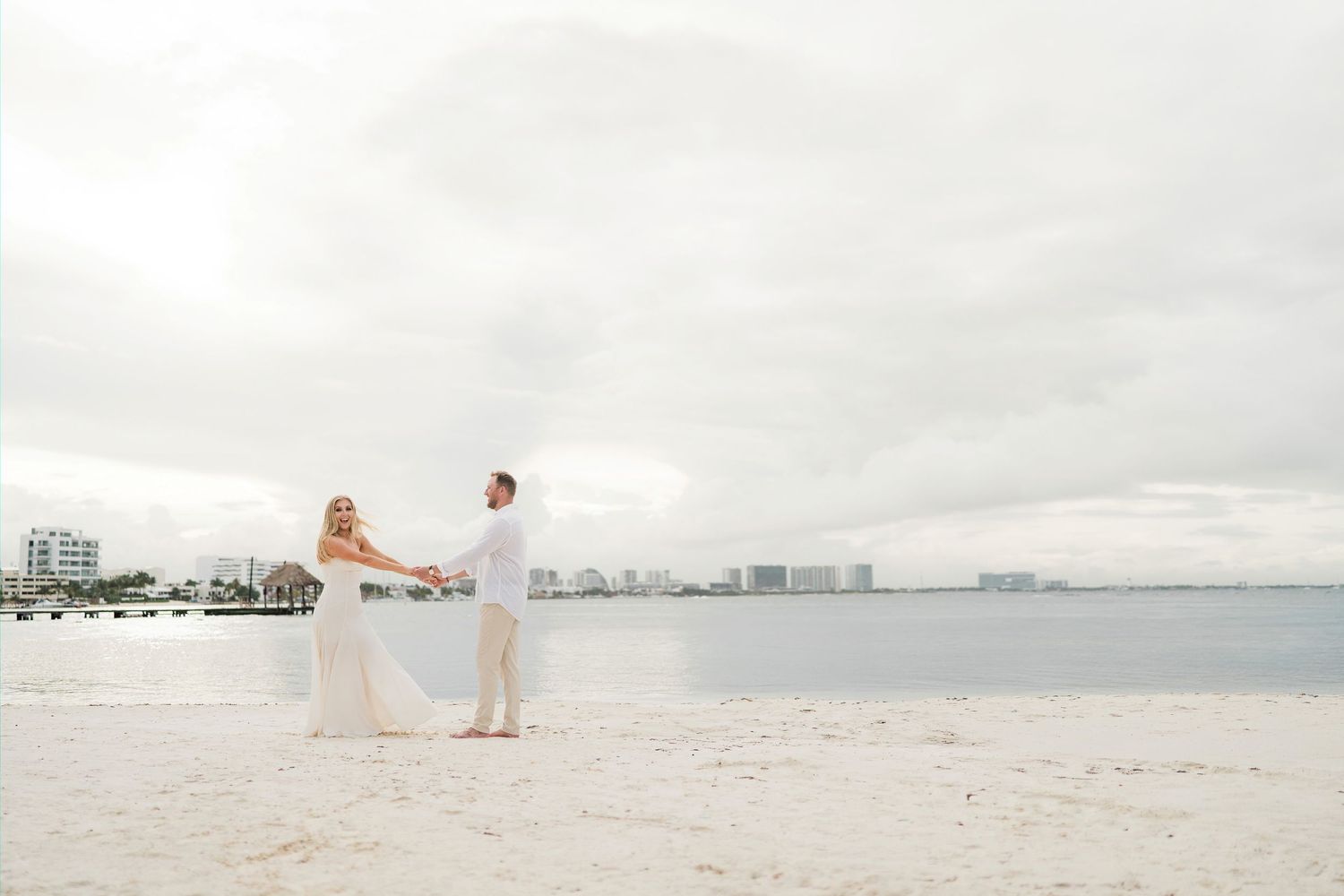 CANCÚN ENGAGEMENT PHOTOSHOOT - Couple holding hands on a white sand beach with ocean views and city skyline under cloudy skies.