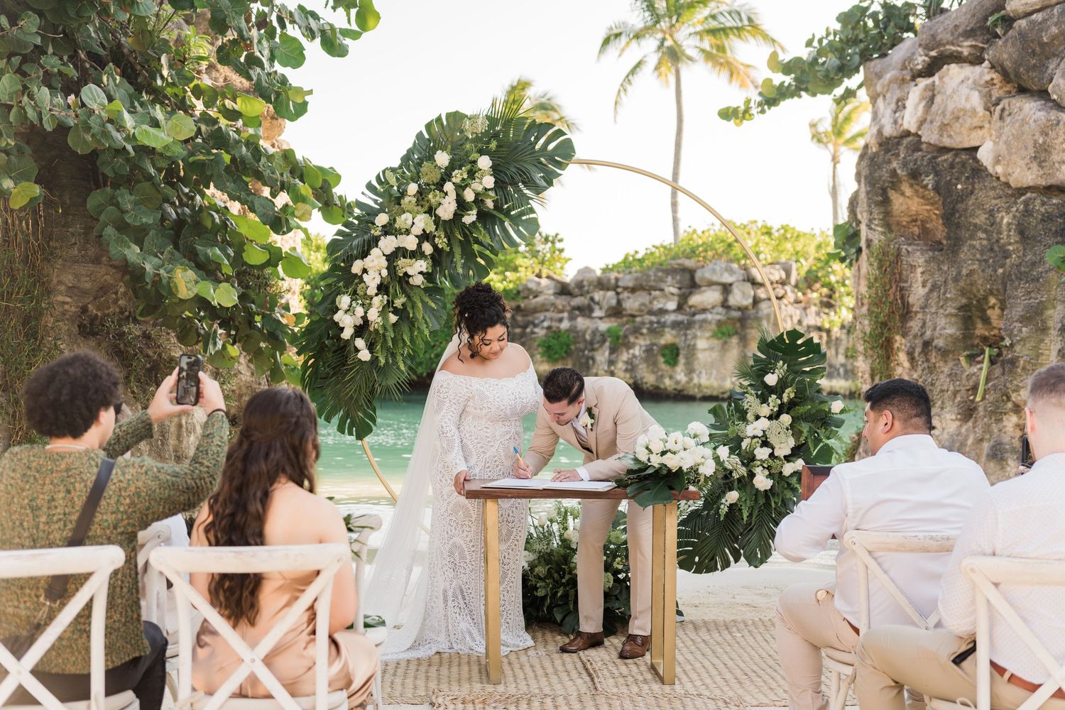 Xcaret Arte Wedding - Couple signing wedding documents at tropical outdoor ceremony with floral arch and turquoise water backdrop.