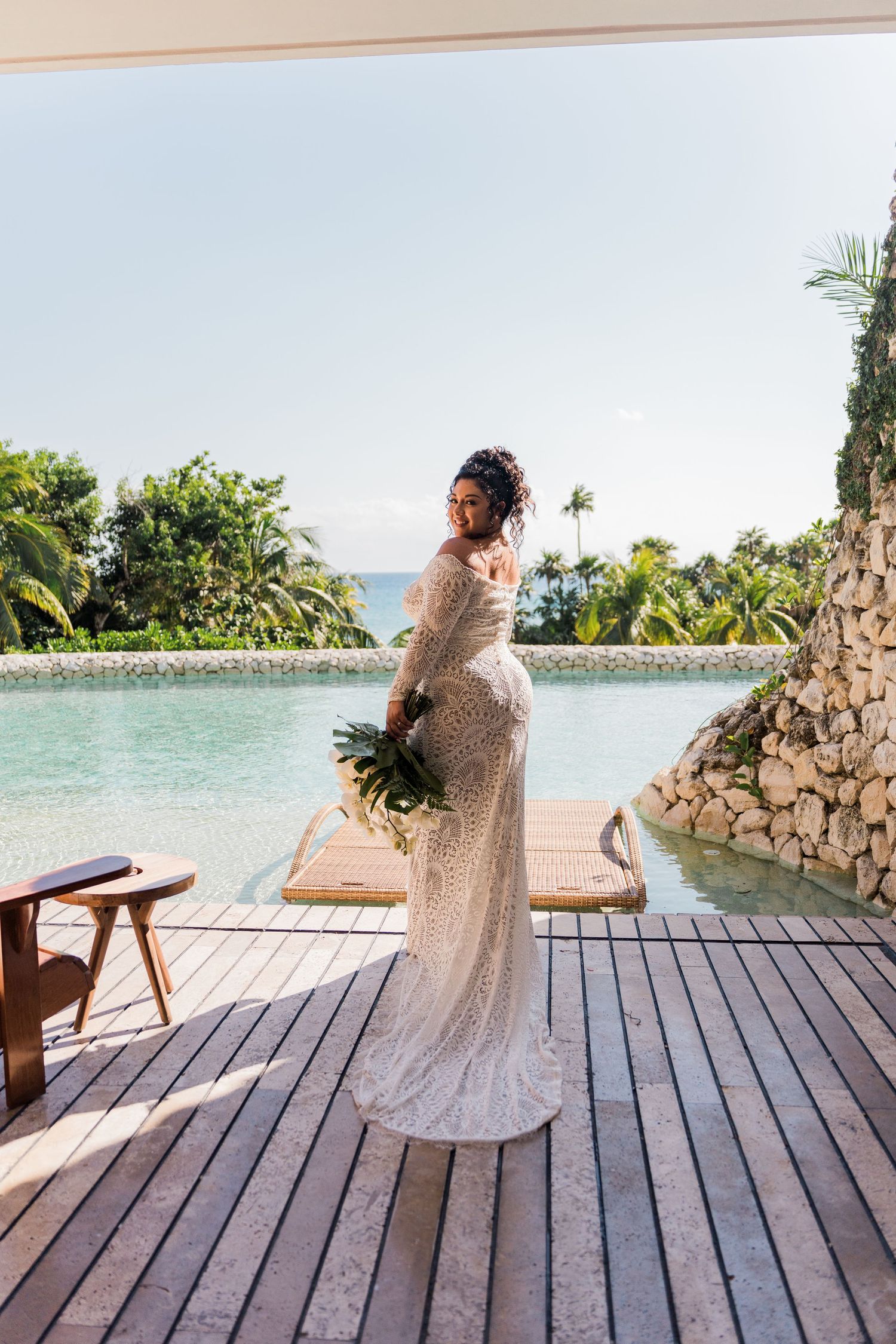 Xcaret Arte Wedding - Bride in lace wedding gown holding bouquet on wooden deck by tropical pool with palm trees and ocean view.