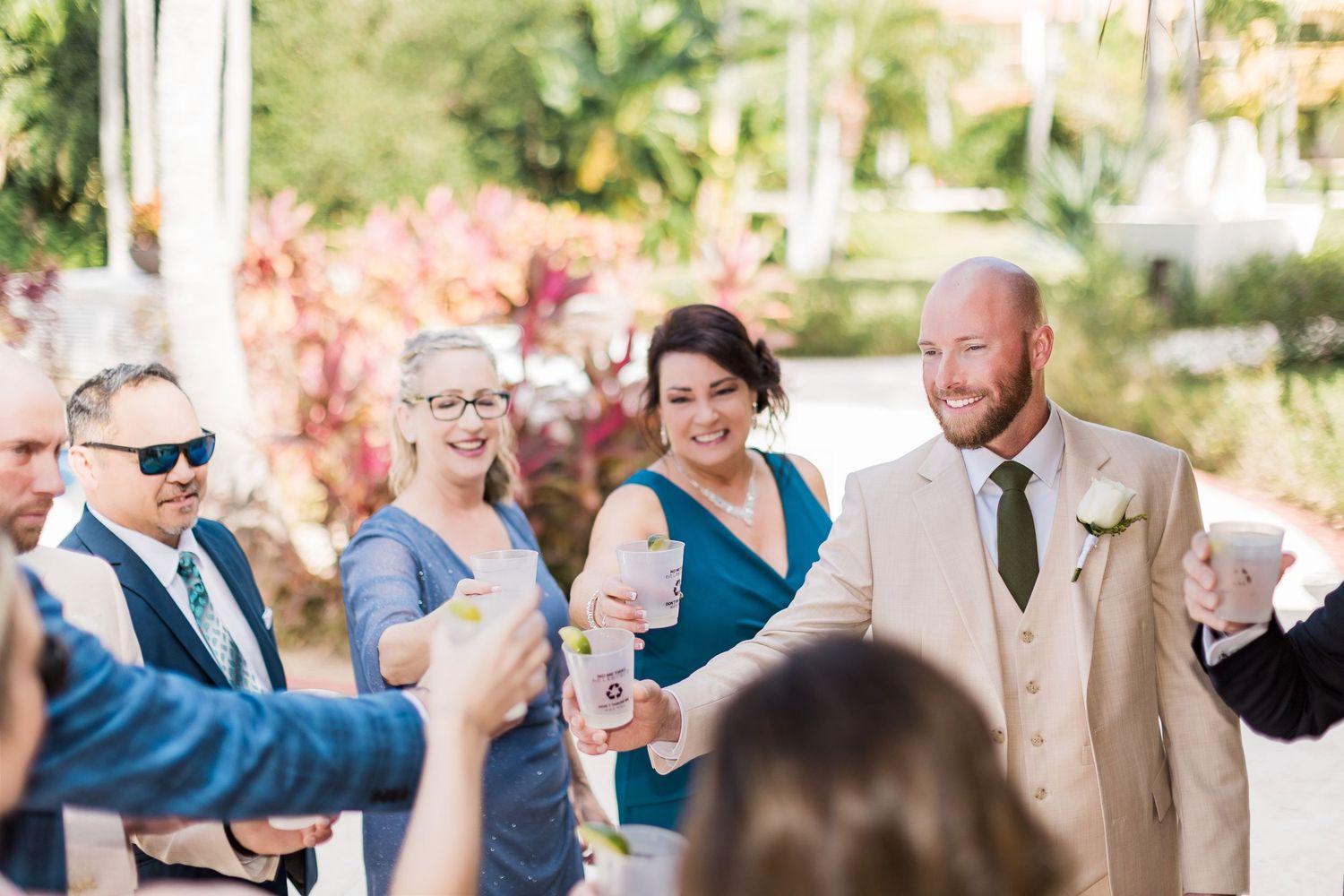 Smiling wedding guests celebrate outdoors with drinks at a sunny tropical venue, bride and groom beaming with joy.
