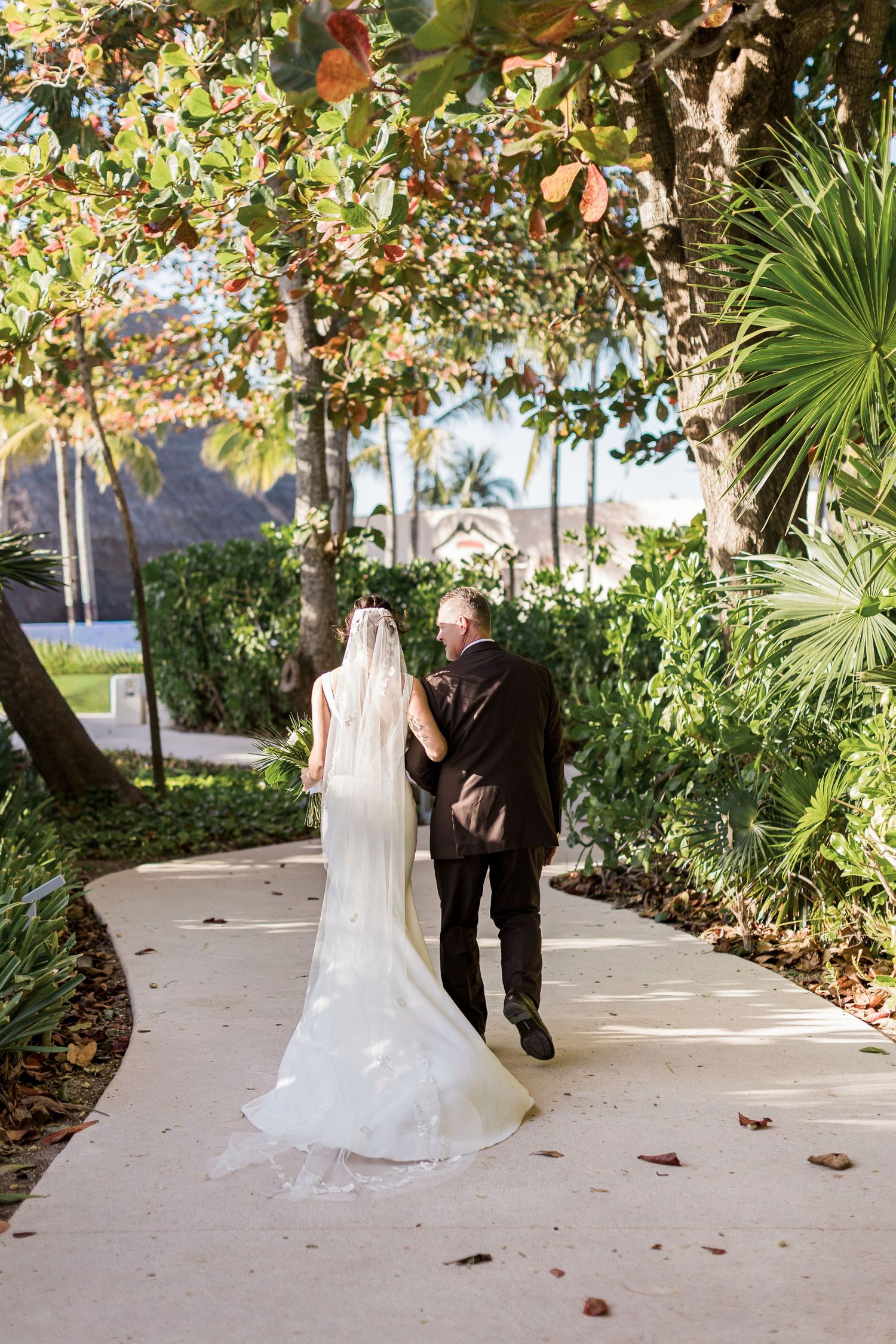 Bride in white gown and veil walks with escort along tropical garden path on wedding day.