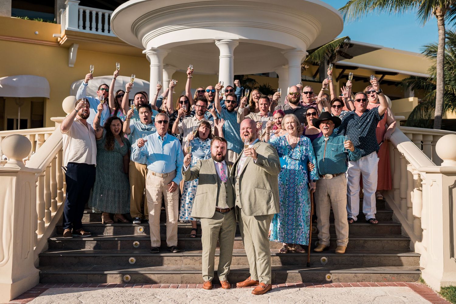 Large group of cheerful people posing on steps of elegant building with white columns at outdoor celebration.