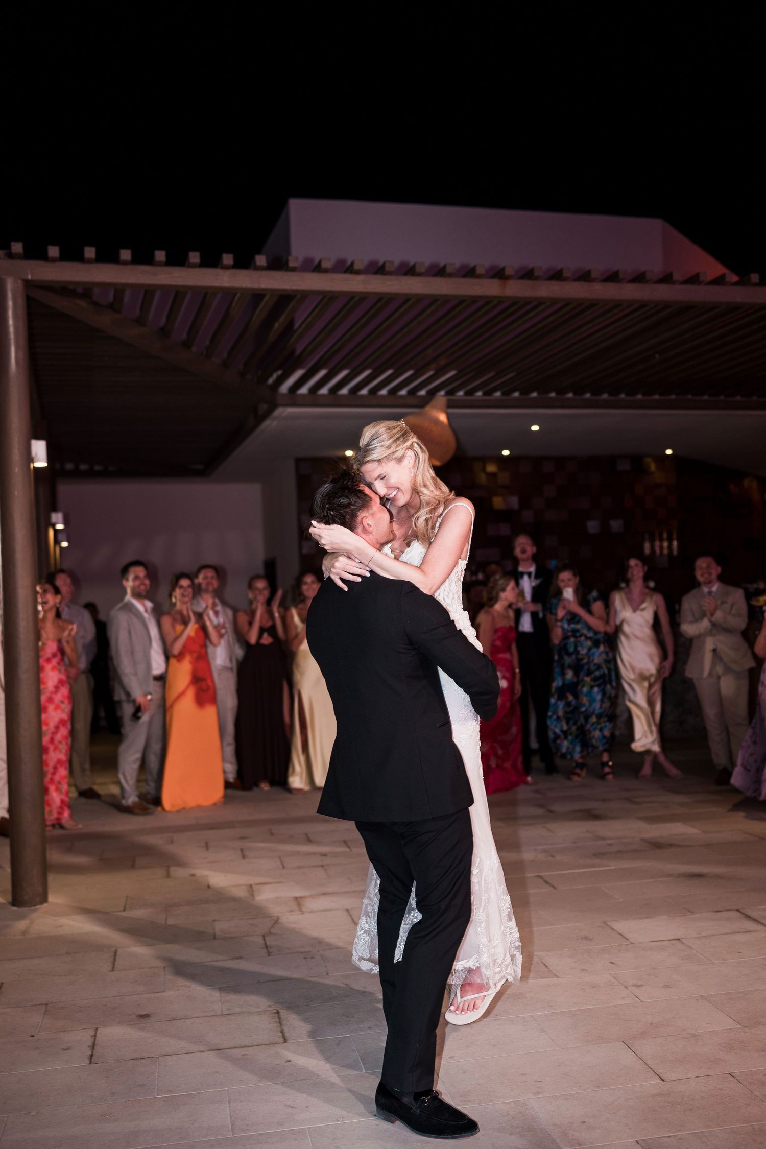 Bride and groom share first dance as groom lifts bride at outdoor wedding reception at night.