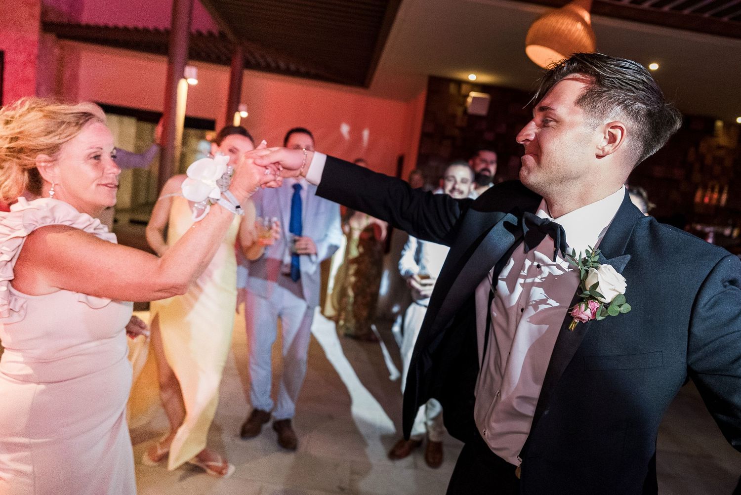 Bride and groom dancing joyfully at wedding reception, groom in tuxedo with boutonniere, guests celebrating in background.