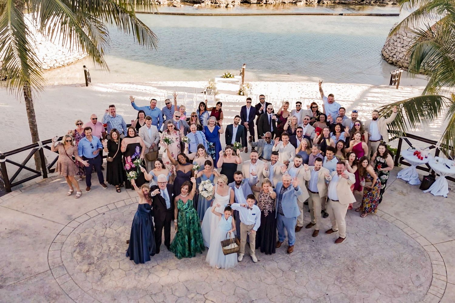 Large wedding group photo outdoors near a tropical beach with palm trees, guests dressed in formal attire celebrating.