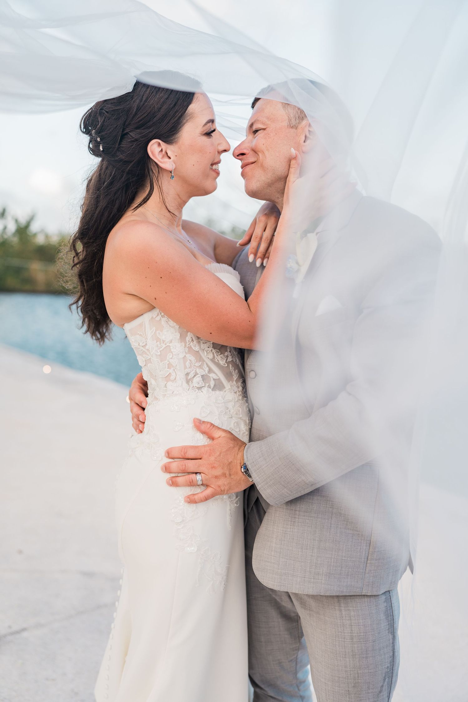 Bride and groom sharing a romantic moment under a flowing veil outdoors near a pool on their wedding day.