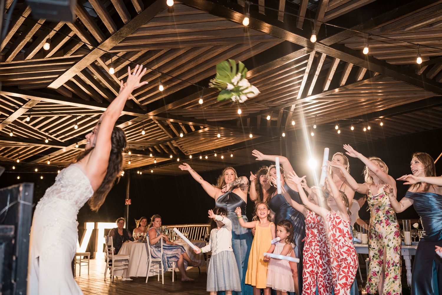 Bride tossing bouquet to excited guests at indoor wedding reception with string lights and wooden ceiling.