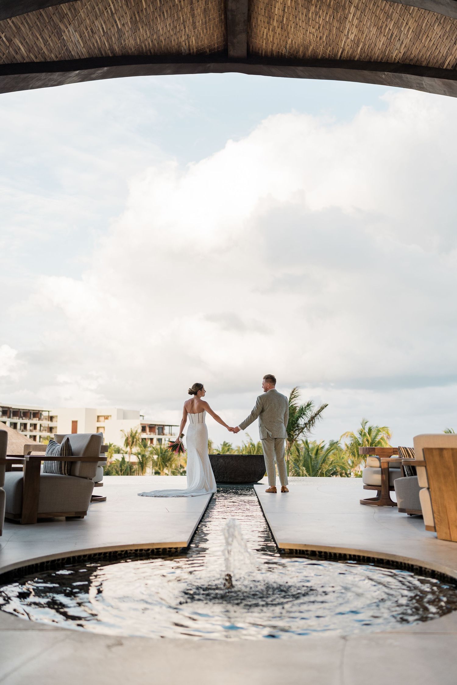Bride and groom hold hands by a reflecting pool at a luxury resort wedding venue with tropical views.
