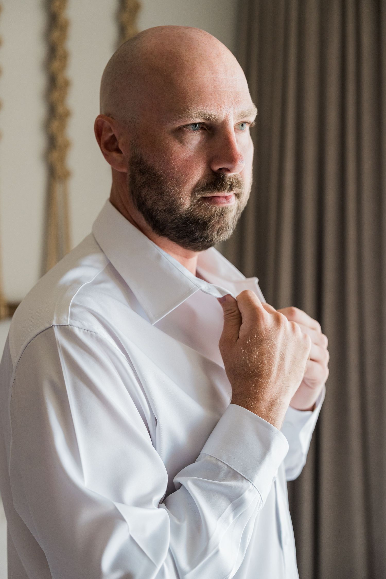 Bald man with beard adjusting his white dress shirt collar, preparing for a formal event.