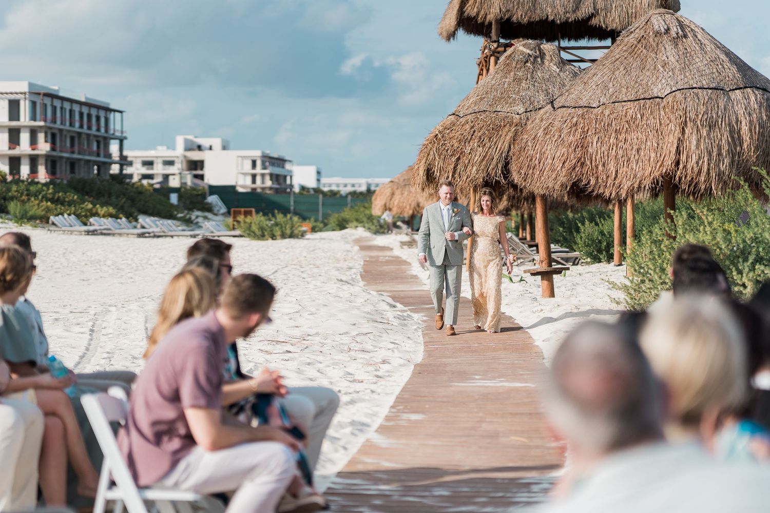 Couple walks down beachside aisle at outdoor wedding ceremony with thatched palapas and guests seated on white chairs.