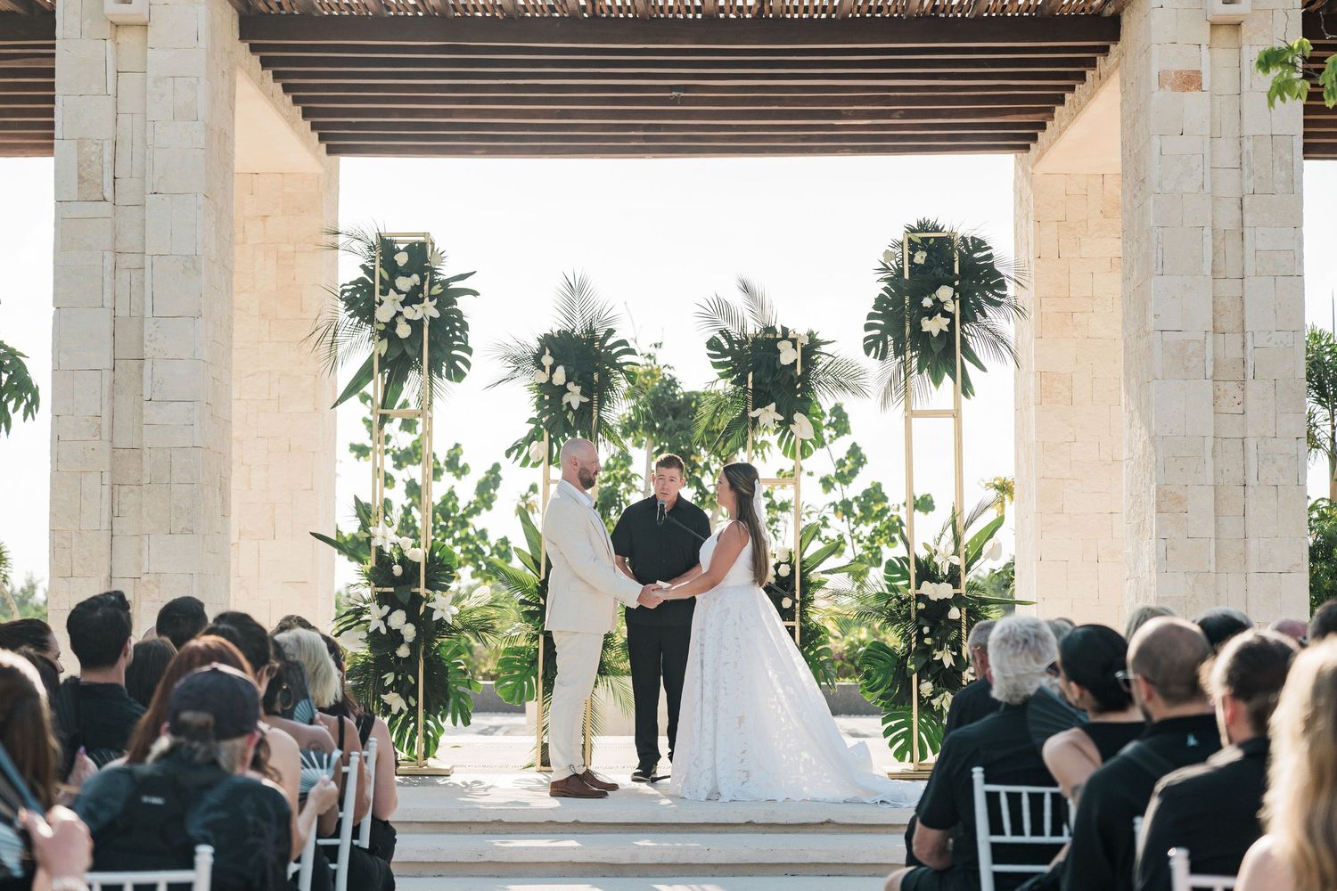 Couple exchanges vows at elegant outdoor wedding ceremony with tropical greenery and white chairs under a pergola.