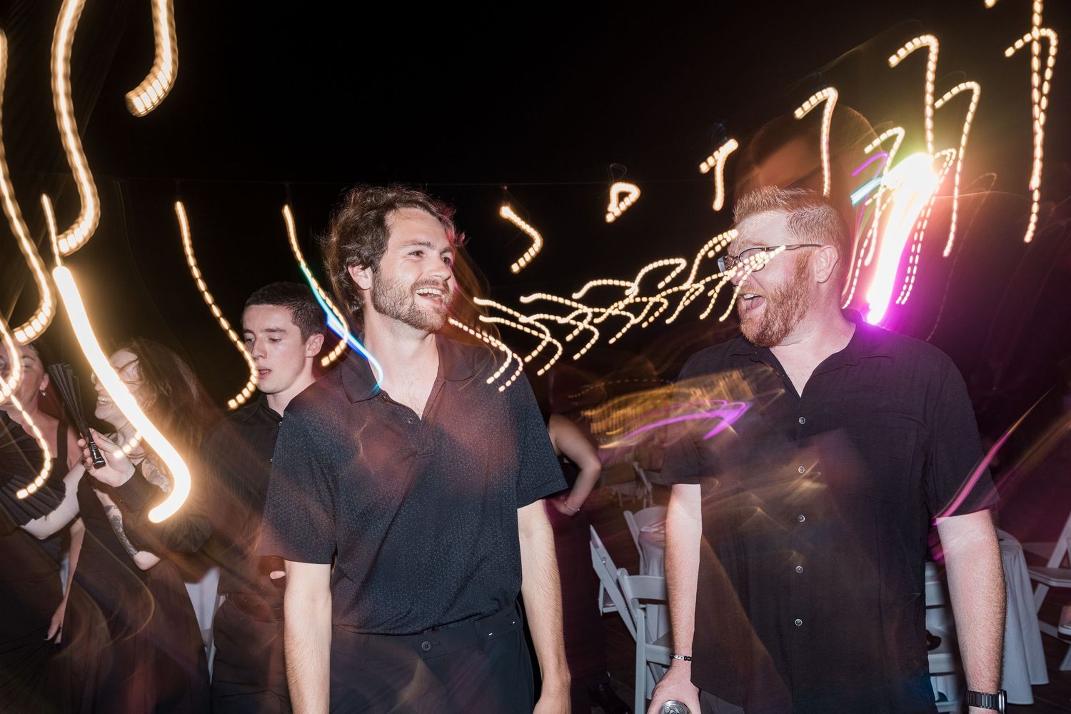 Two men smiling at a nighttime outdoor event with colorful light trails in the background.