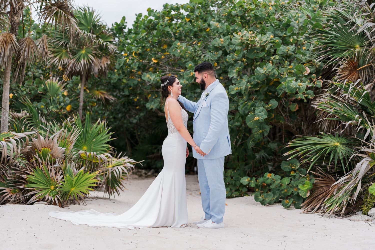 Bride and groom holding hands facing each other on a white sand beach with lush greenery.