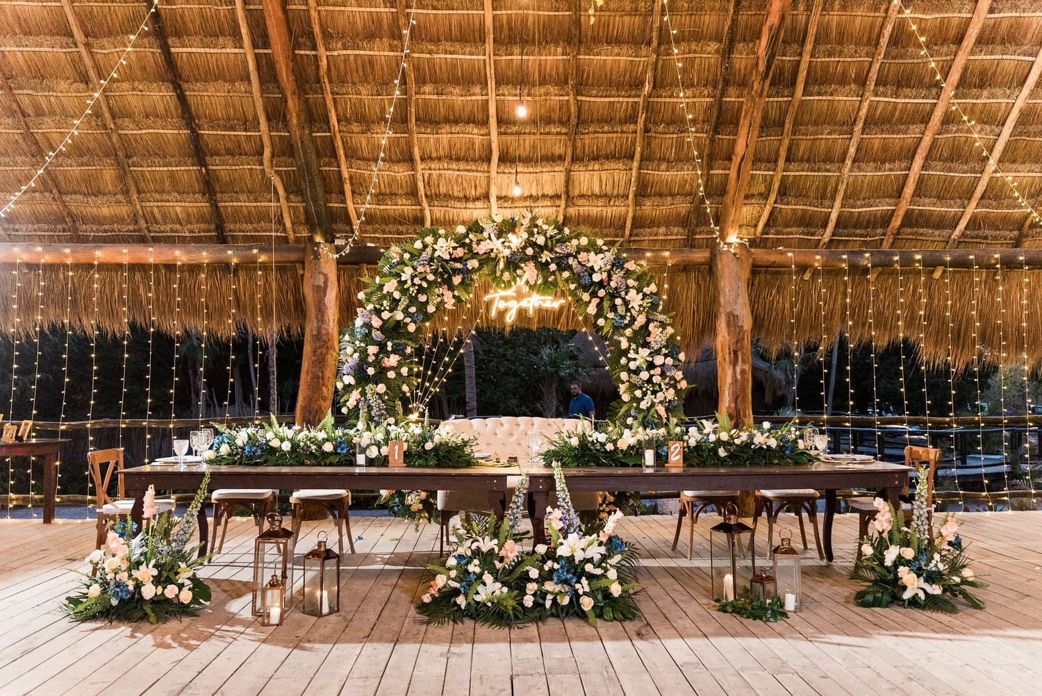 Elegant indoor wedding reception with floral arch, string lights, and long decorated tables.