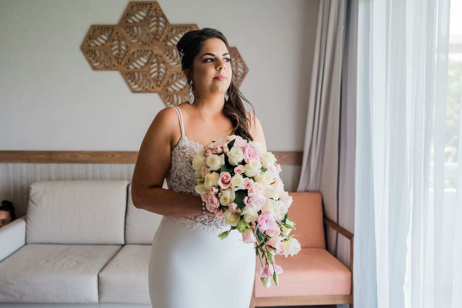 Elegant bride in beaded gown holding pink and white cascading bouquet by hotel window.