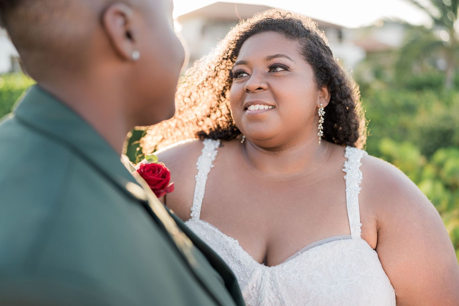 Bride in white lace dress smiles lovingly at partner in green suit with red boutonniere at outdoor wedding.