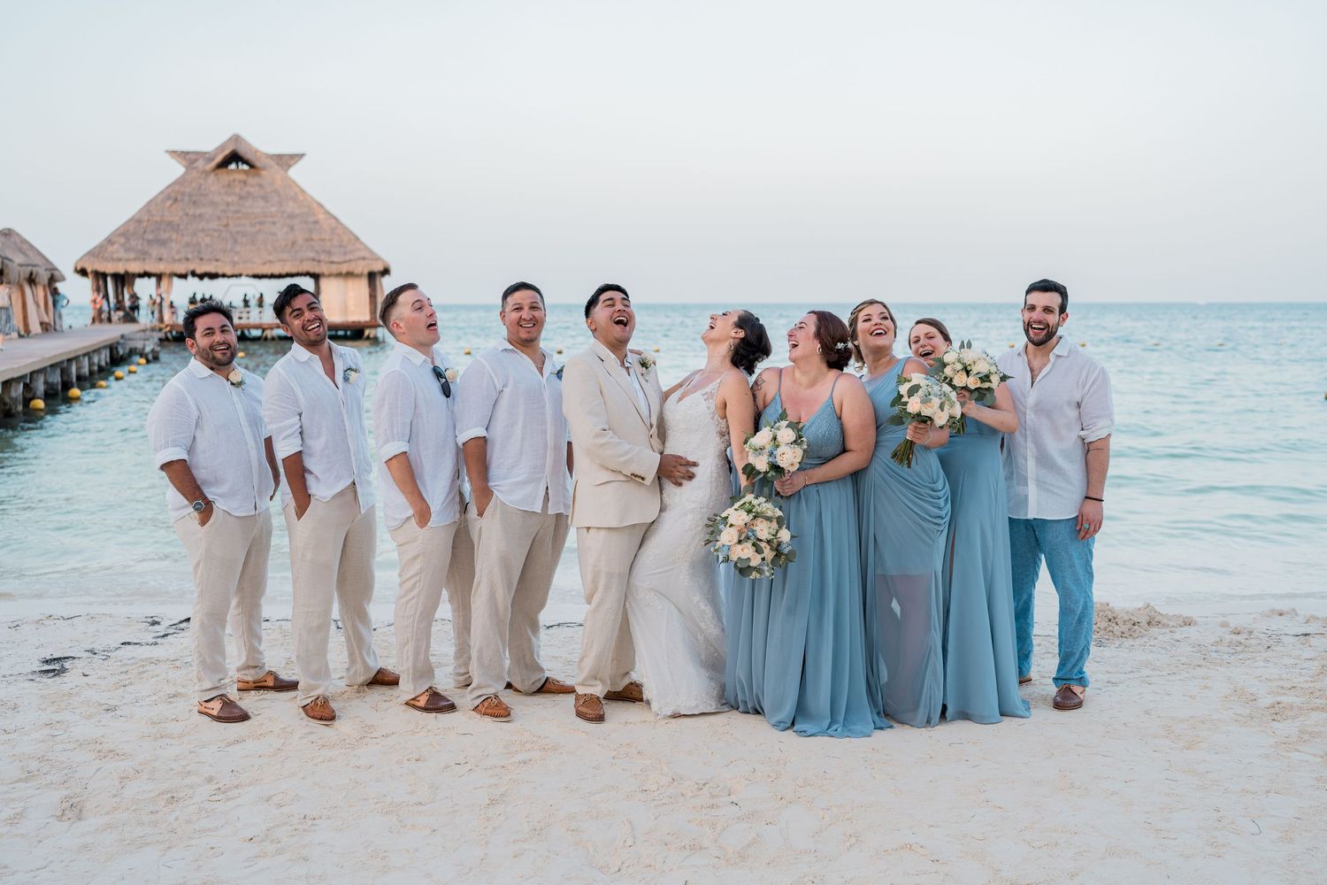 Bridal party in dusty blue and beige poses playfully on Caribbean beach at dusk.