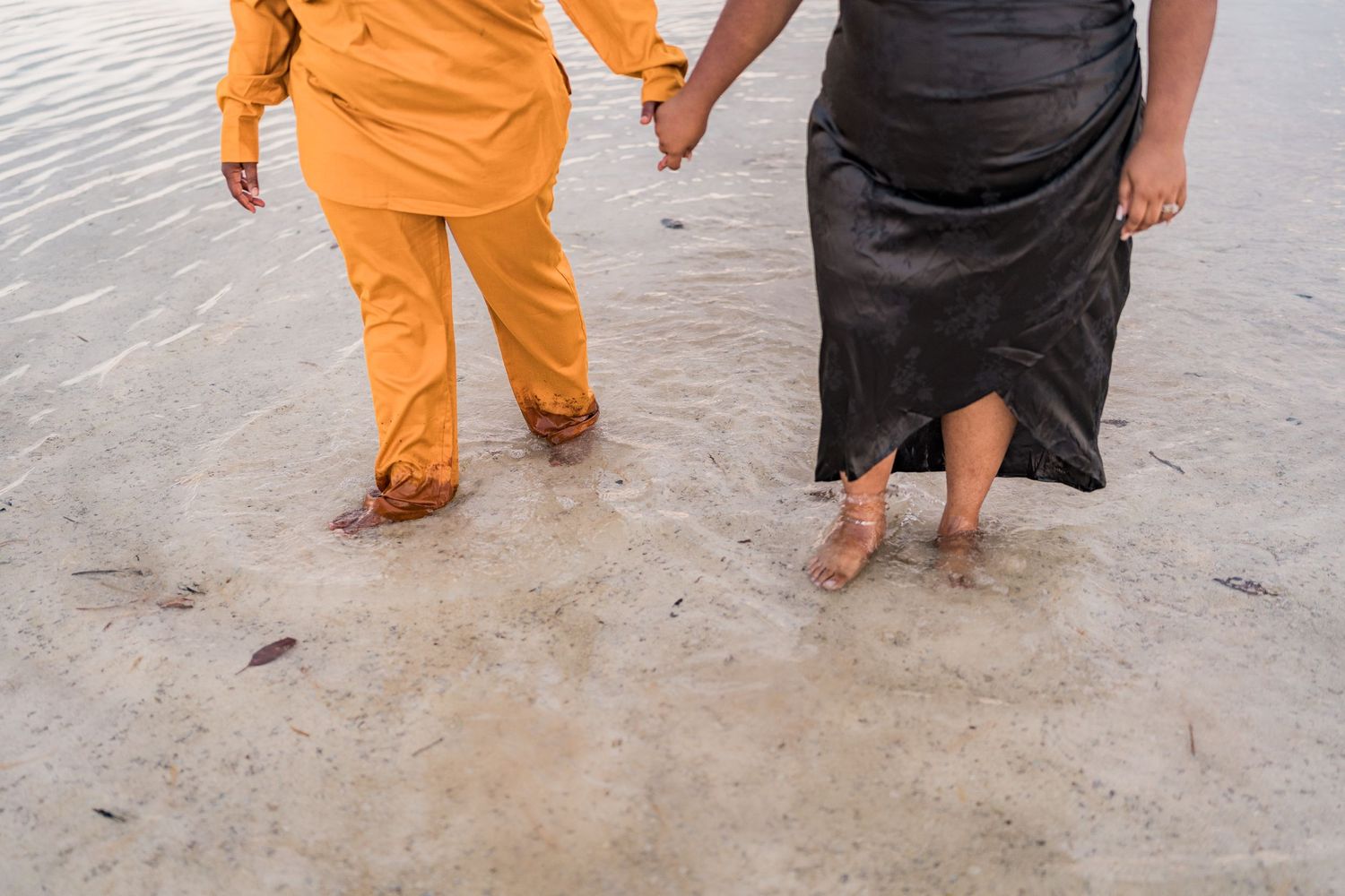 Couple holding hands walking barefoot in ocean waves, one wearing orange outfit and other in black dress.