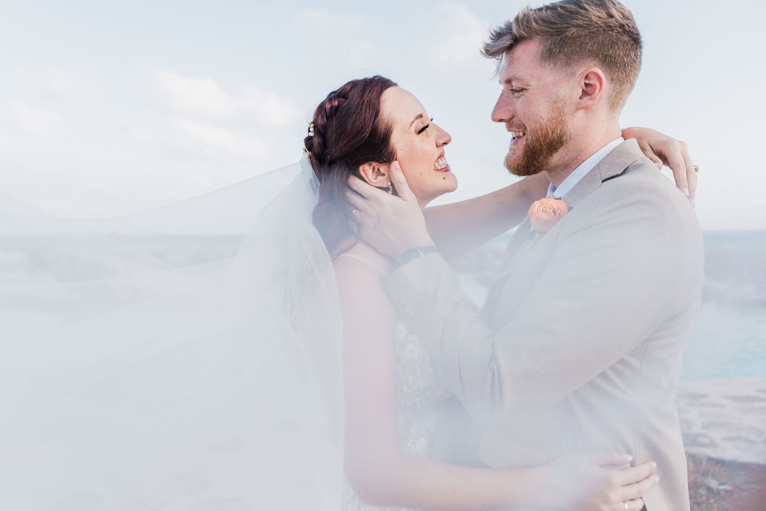 Bride and groom embrace romantically under flowing veil with misty ocean backdrop at beach wedding.