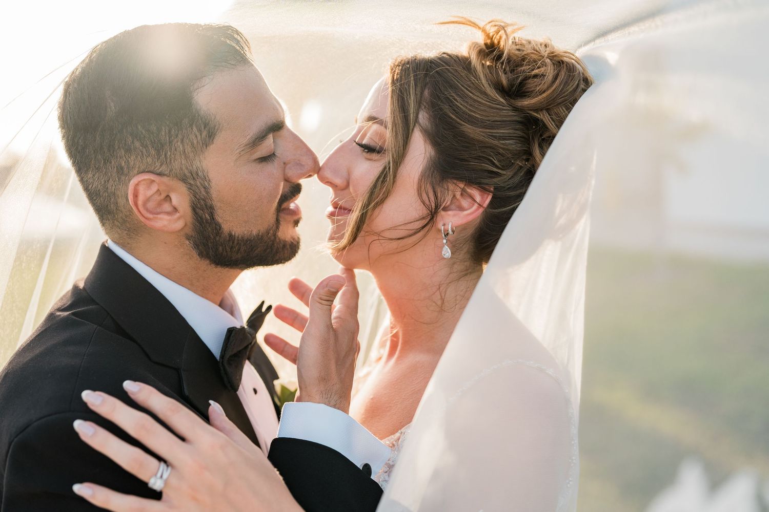 Bride and groom share a tender almost-kiss under a flowing veil in golden sunlight.