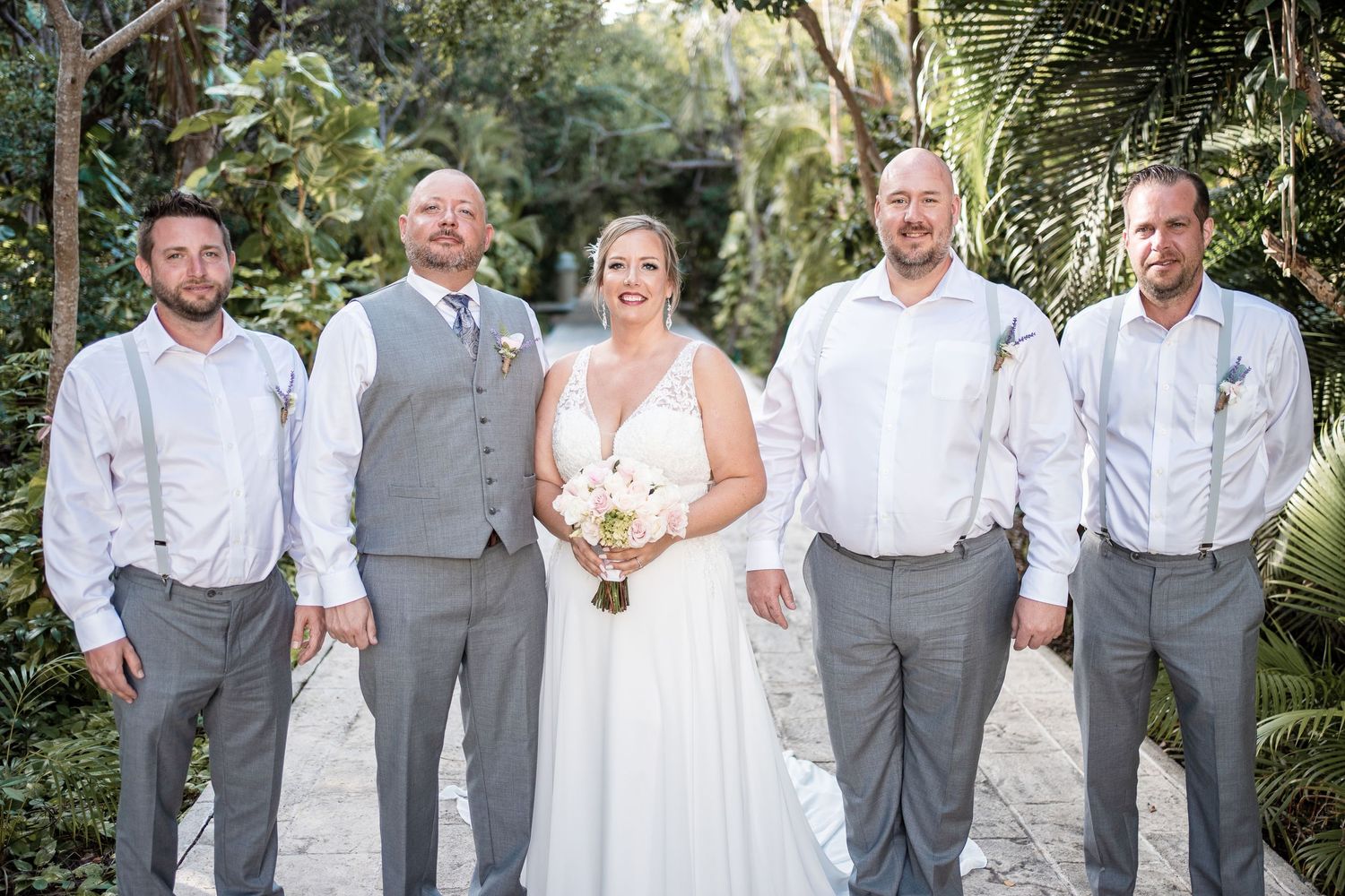 Bride with bouquet poses with four groomsmen in gray pants on tropical garden pathway.