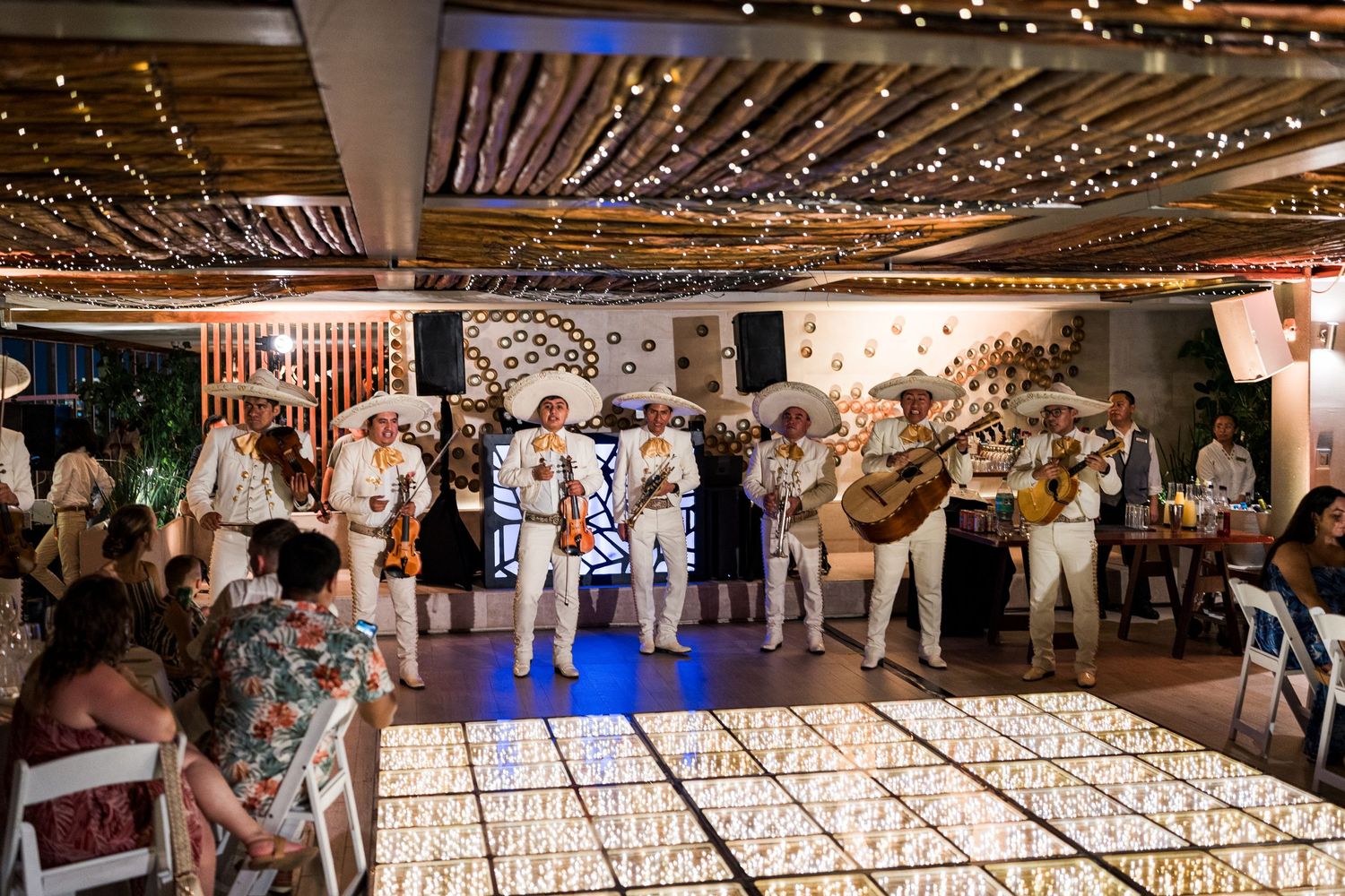 Mariachi band performs on a lit dance floor at an outdoor beach wedding reception at night.