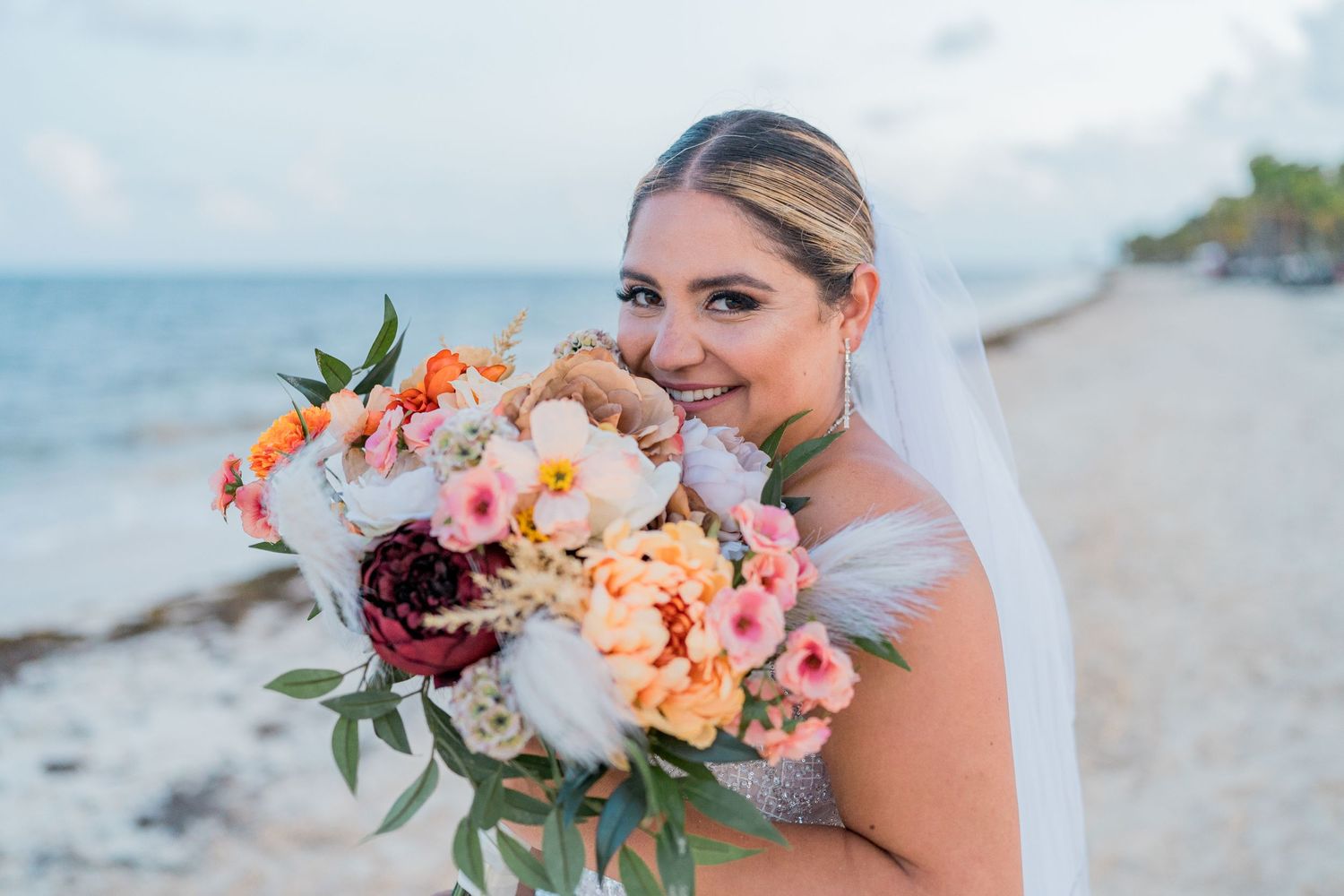 Radiant bride holding a colorful bouquet of pink, red, and orange flowers on a beach wedding day.