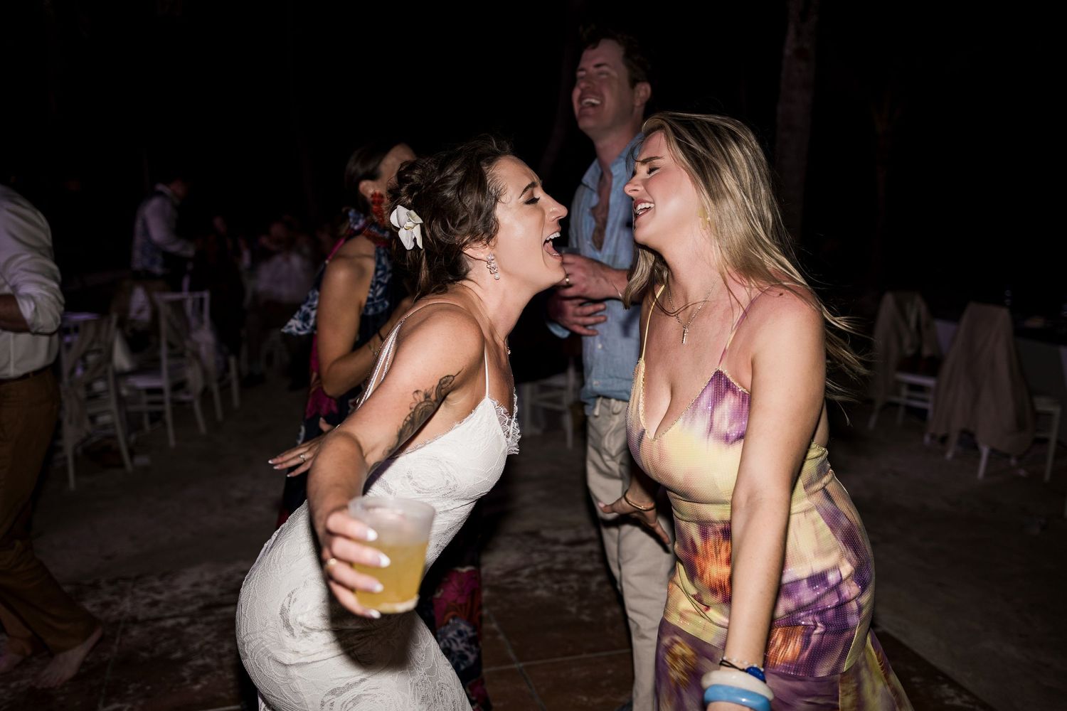 Two women laugh and dance together at an outdoor evening wedding reception, one in white bridal gown holding a drink.