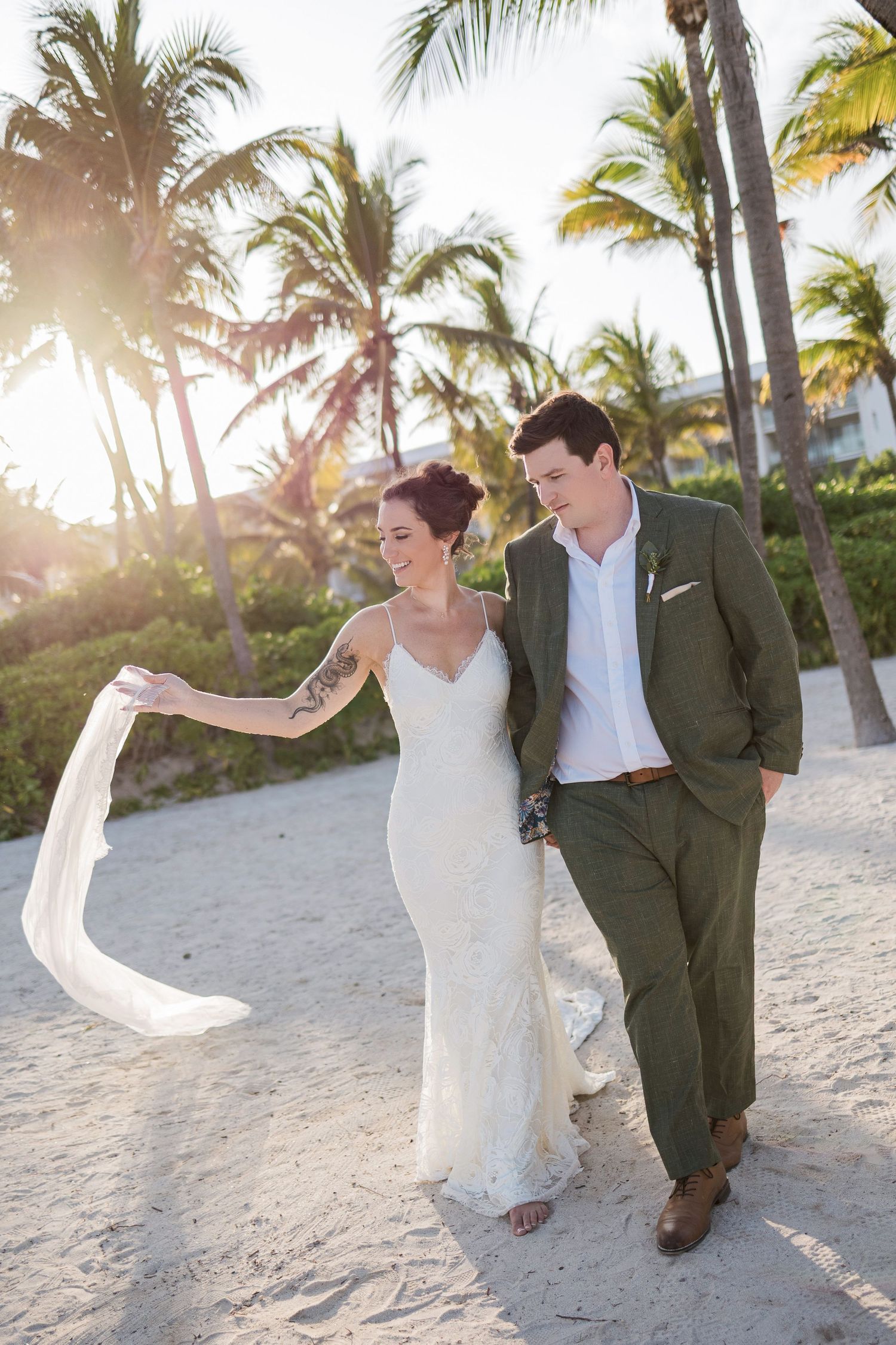 Bride and groom walking on sandy beach at sunset, palm trees, white wedding dress with veil, green suit.