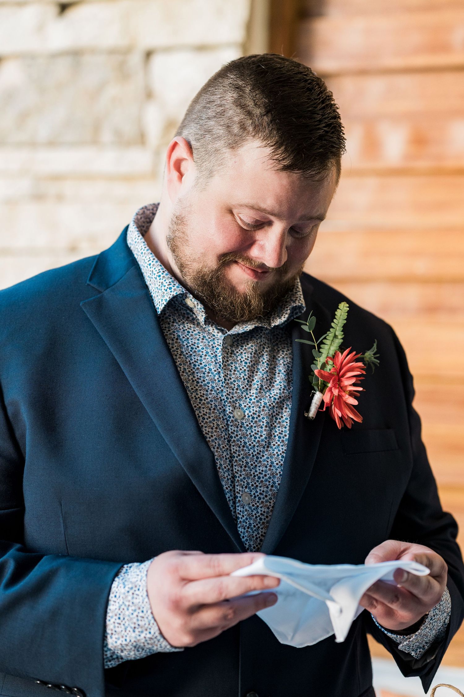 Secrets Akumal Wedding - Groom in navy suit with red floral boutonniere reads a note, smiling softly at a rustic wedding venue.