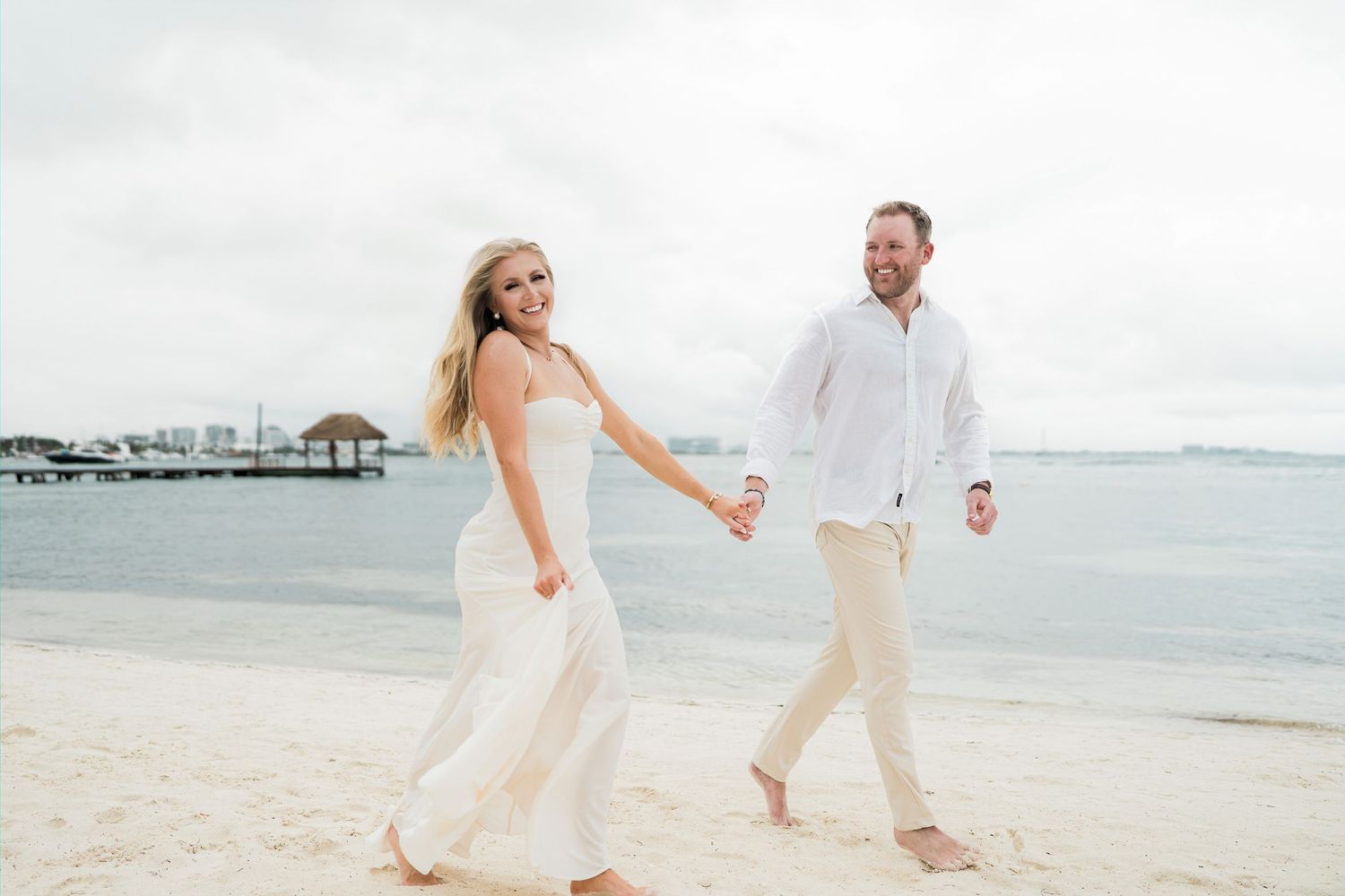 CANCÚN ENGAGEMENT PHOTOSHOOT - Happy couple in white attire holding hands and walking on a tropical beach near calm ocean waters.