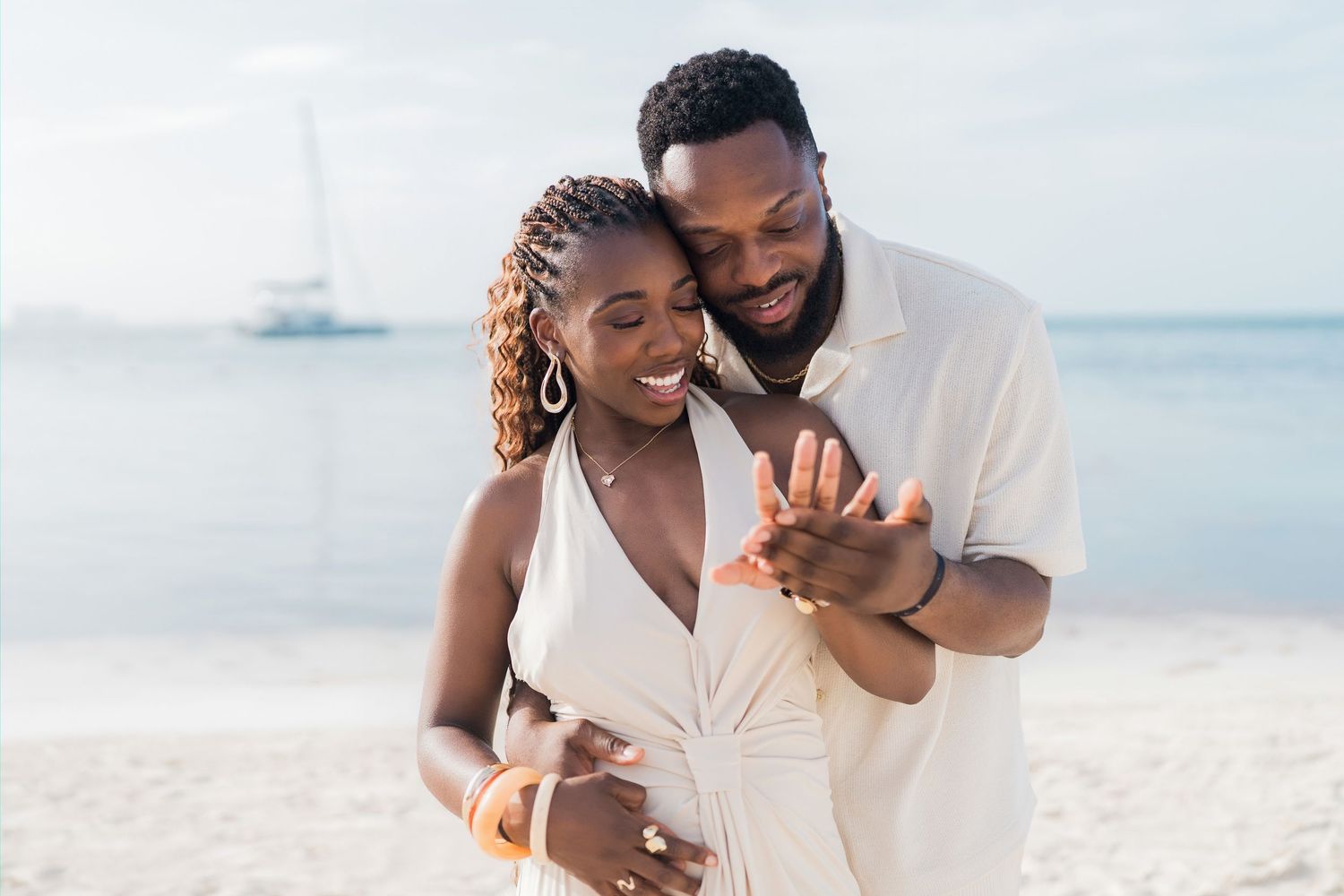 Happy couple on beach, woman in white dress admiring engagement ring, sailboat in background.