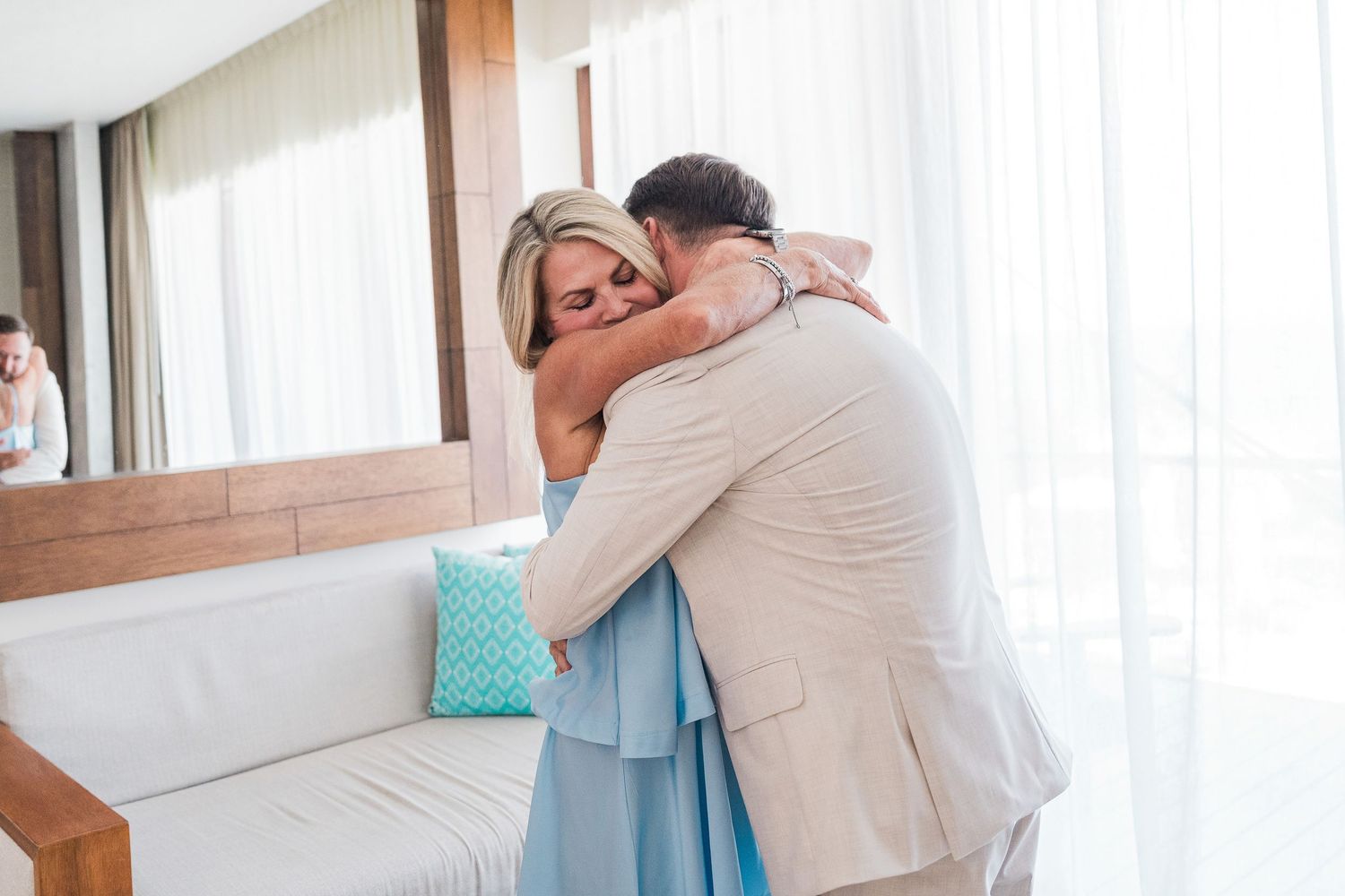 A couple shares a warm embrace in a bright room, the woman wearing a blue dress hugging a man in a beige suit.
